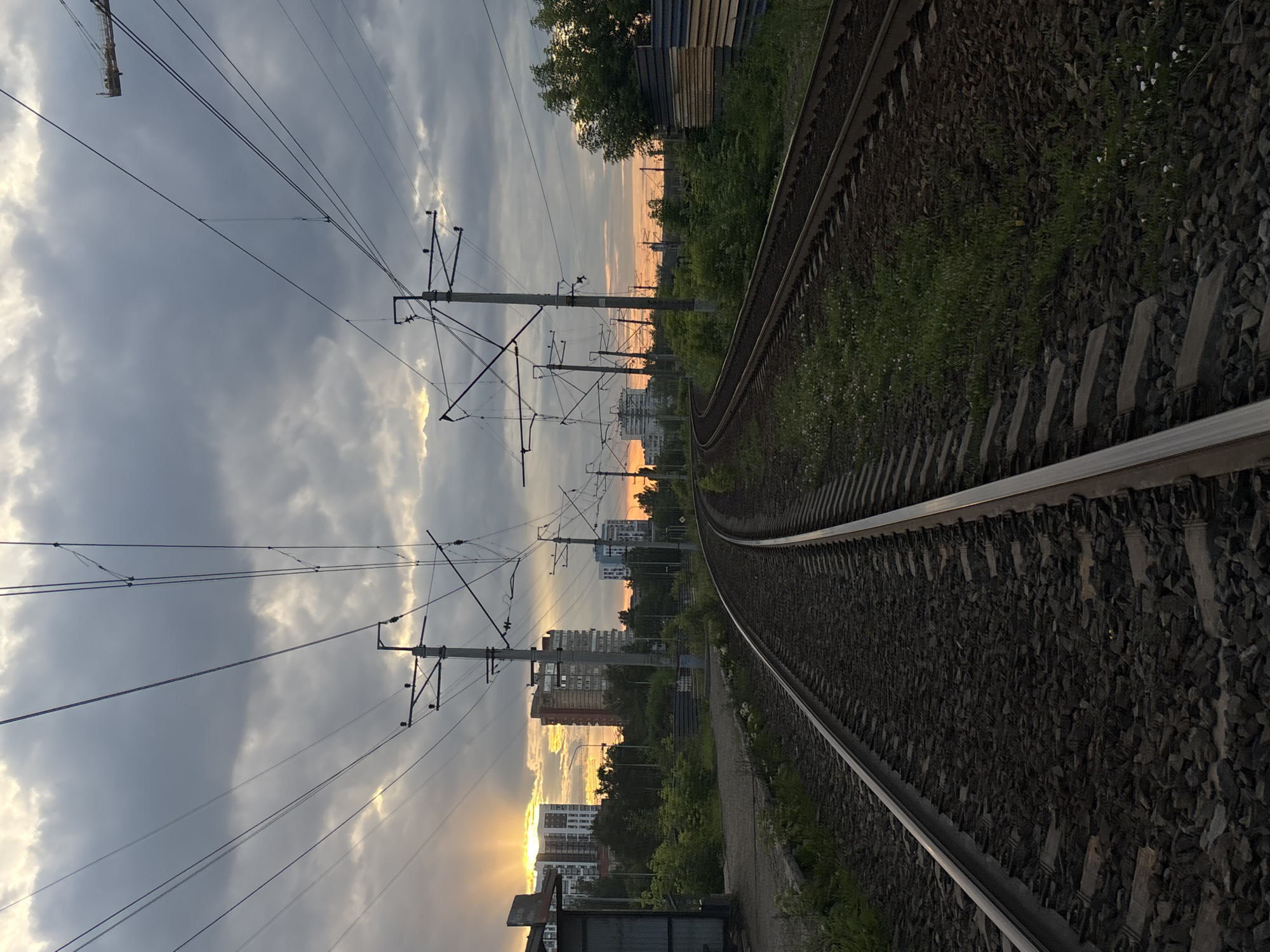The image depicts a view of a railway track in Perm, Russia, under a cloudy sky at sunset. The railway tracks curve to the left in the foreground, with a second set of tracks visible to the right. Overhead, a complex network of power lines and support poles stretches across the frame. In the background, several multi-story buildings are visible, suggesting an urban setting. The sun is setting behind some of these buildings, casting a warm glow and rays of light through the clouds. Green vegetation, including trees and bushes, borders the railway and the urban structures. A small, partially visible building, possibly a bus stop or shelter, is located on the left side of the frame near the tracks. The scene is devoid of any people or visible action.