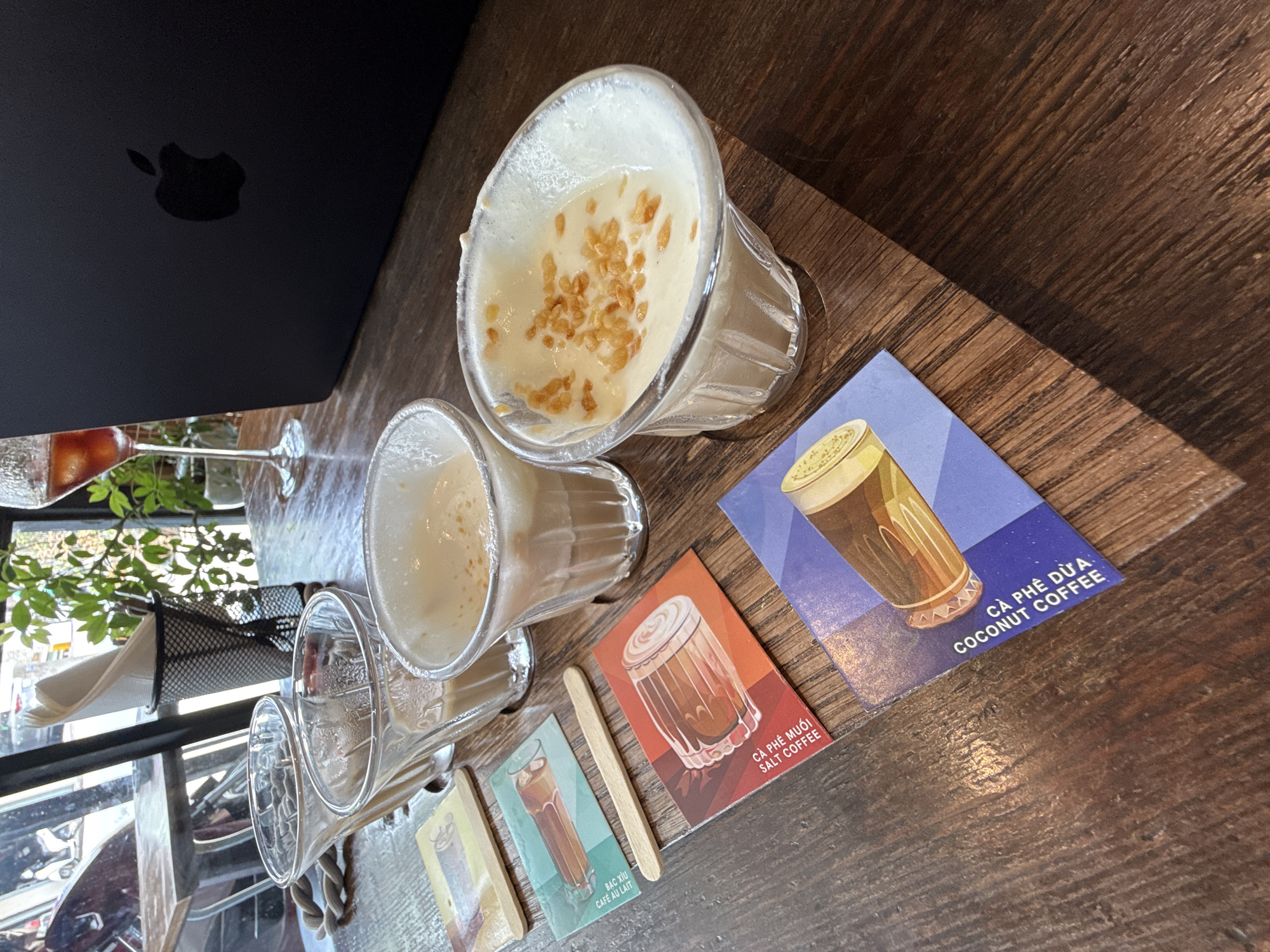 The scene depicts a variety of Vietnamese coffees served in clear glass containers atop a wooden table, alongside informational cards detailing each coffee type, within what appears to be a cafe setting. Dominating the scene are two partially-consumed coffee drinks in clear glass vessels; one with a white foamy top sprinkled with crunchy elements. Alongside these are smaller glasses, either empty or containing remnants of coffee. Several small cards display illustrations of the coffees they represent. The cards are placed in a row, indicating different coffee options available at the establishment. The card descriptions include "Cà Phê Dừa Coconut Coffee," "Cà Phê Muối Salt Coffee," and "Bạc Xỉu Café Au Lait." To the left, a black laptop with an Apple logo is present. A table and window with greenery can be seen to the side. The photograph was taken during daylight hours, indicated by the natural light visible through the window. Based on the types of coffee served and the visible location, it is implied the setting is Da Lat, Vietnam.