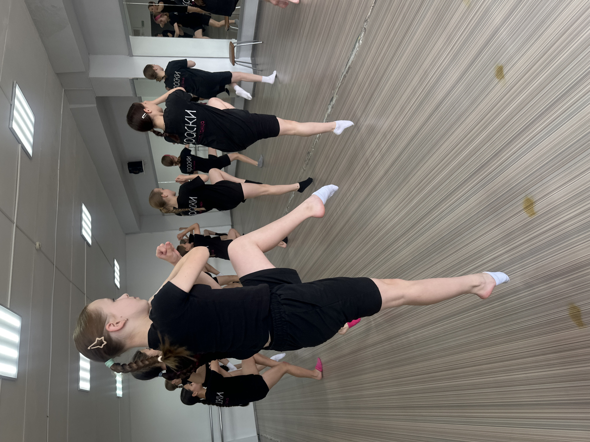 The image depicts a dance studio, likely located in Votkinsk, Russia, where a group of young girls are engaged in a dance class. The room features a mirrored wall, a barre, and bright fluorescent lighting. The girls are all dressed in black t-shirts and shorts, and some are wearing white socks while others have pink slippers. The floor is made of light-colored wood with a striped pattern. One girl in the foreground is captured mid-movement, with her left leg extended upwards in a kick. She has a star-shaped hair clip in her braided hair. The other girls are also in various stages of stretching or movement, facing a mirror. The overall atmosphere suggests a focused and active learning environment.