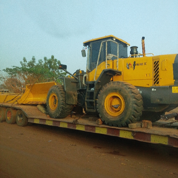 A yellow wheel loader rests on a flatbed trailer with multi-axle wheels. The trailer has a weathered appearance with yellow and red stripes. The loader's bucket is facing forward. The machinery is in a slightly dirty state, indicating active use. Background greenery is visible under a cloudy sky. The ground appears to be reddish dirt. The loader's brand features stylized Chinese characters.