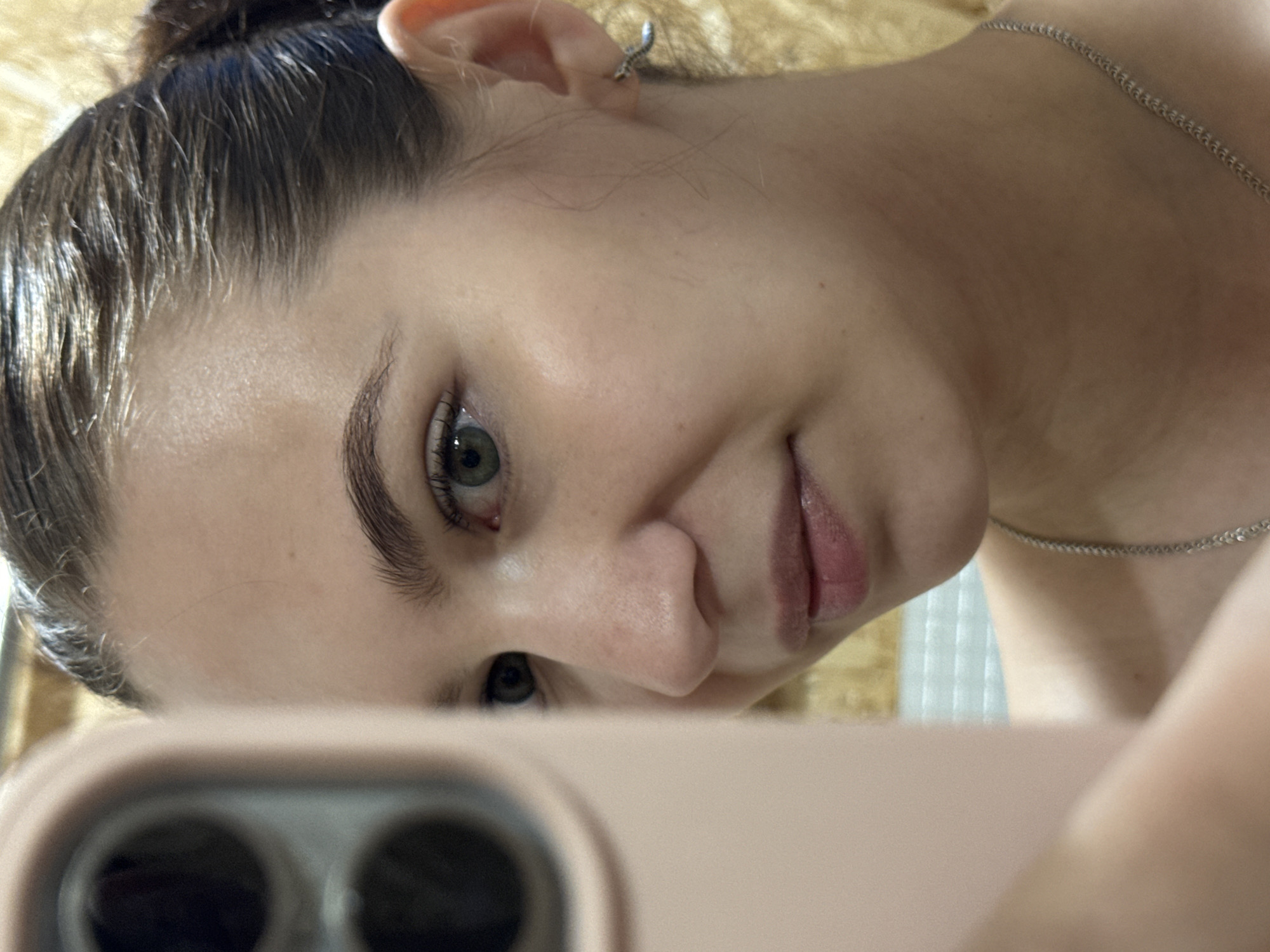 A young woman is taking a selfie, holding her pink smartphone camera up at an angle. She has dark hair pulled back, fair skin, and green eyes. She is wearing a silver chain necklace and a small hoop earring. The background is out of focus. The location is not identifiable from the image.