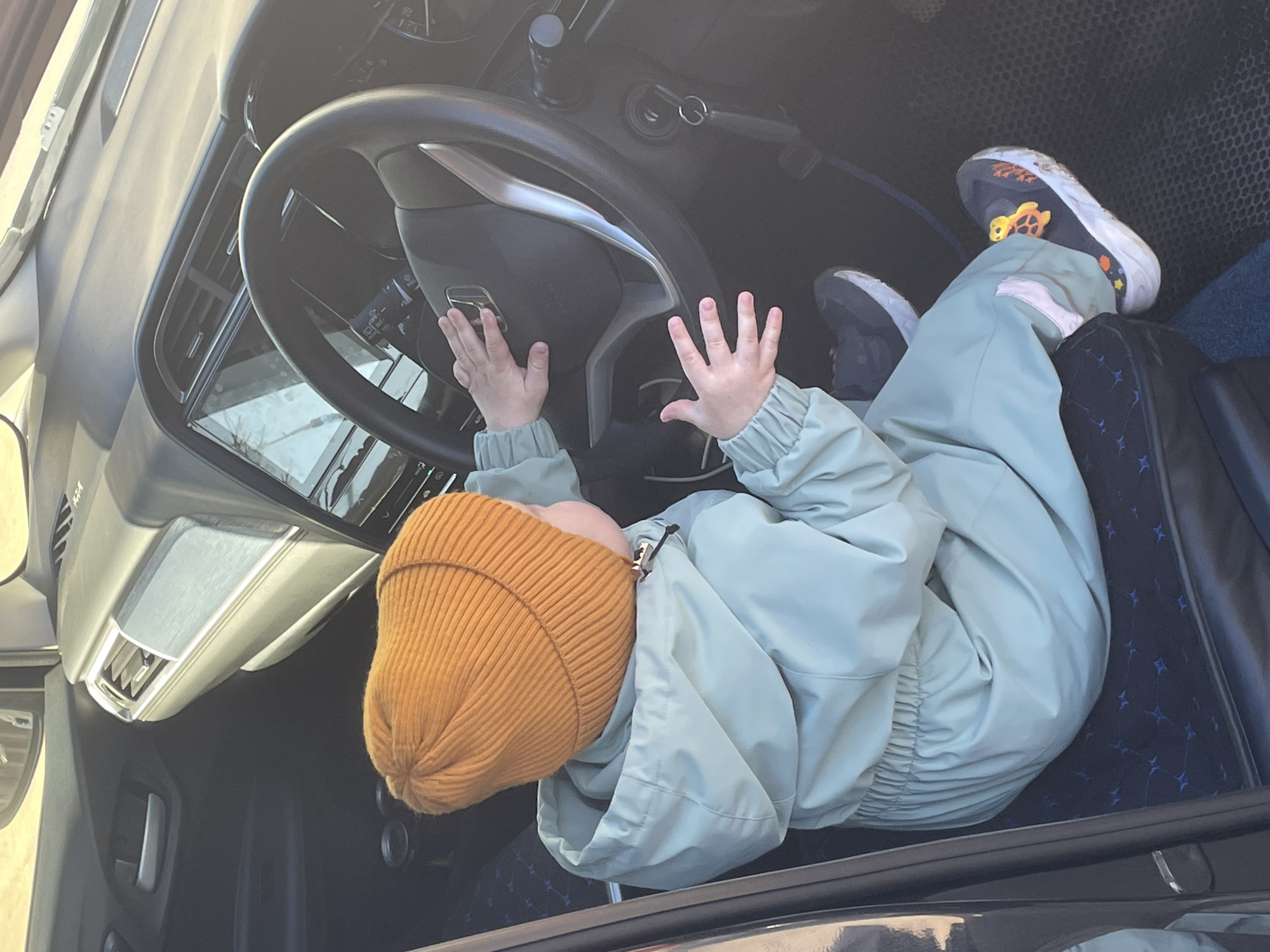 A young child wearing a mustard-yellow knit hat and a light green jacket is seated in the driver's seat of a car. The child's hands are on the steering wheel. The location in the city of Siva, Russia, cannot be determined from the image.