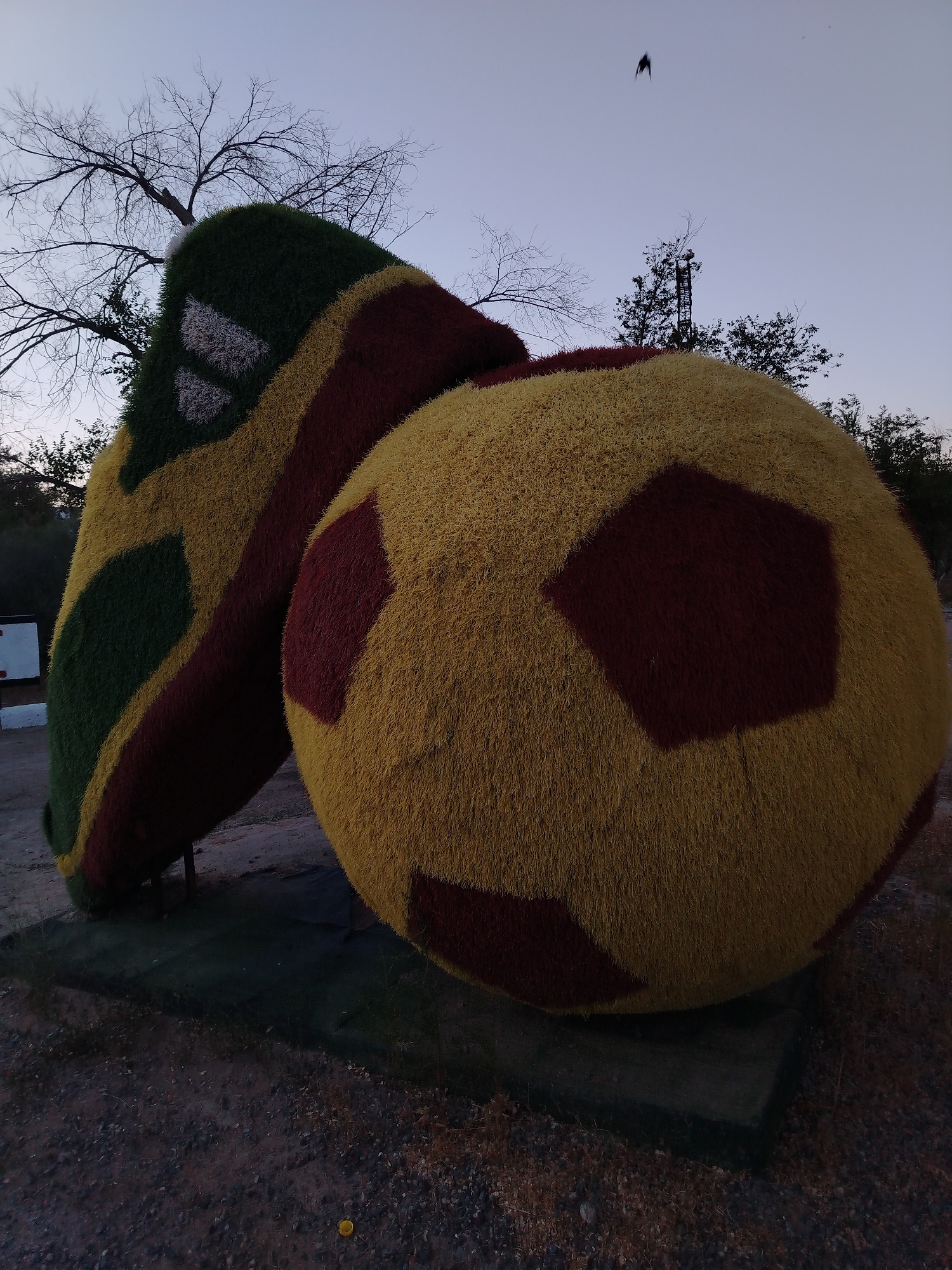 The image shows a large, stylized soccer ball and cleat sculpture covered in artificial grass. The soccer ball is primarily yellow with dark red pentagonal panels, while the cleat is green and yellow with dark red accents and white stripes. The sculpture is located outdoors, on a dirt and gravel ground. In the background, there are bare trees and a clear sky with a hint of dusk. A faint silhouette of a bird is visible in the sky, and a distant radio tower can be seen behind the sculpture. The scene is likely part of a park or public space in Baikonur, Kazakhstan, possibly related to sports or a themed attraction.