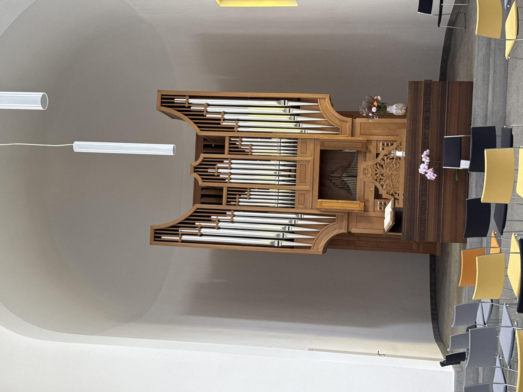 The image depicts the interior of a modern church sanctuary, likely in Sesto San Giovanni, Italy, characterized by its minimalist design and architectural features. Dominating the scene is a large, wooden pipe organ with gleaming silver pipes arranged in a decorative manner. The organ is situated on a raised platform behind a wooden altar or communion table. On the altar, a book is open, and a vase of flowers, along with a glass of water, are present. The wall behind the organ features a subtle architectural curve, and two cylindrical fluorescent light fixtures hang from the ceiling.
In the foreground, rows of chairs are arranged on a tiered floor. The chairs vary in color, with some being grey, yellow, orange, and black. The floor is tiled in a neutral tone. The overall lighting is soft, casting gentle shadows. The environment appears to be clean and well-maintained, suggesting an active place of worship. The architectural style is contemporary, with smooth, curved surfaces and clean lines. The limited number of visible people, primarily implied by the presence of the chairs, suggests a moment before or after a service.