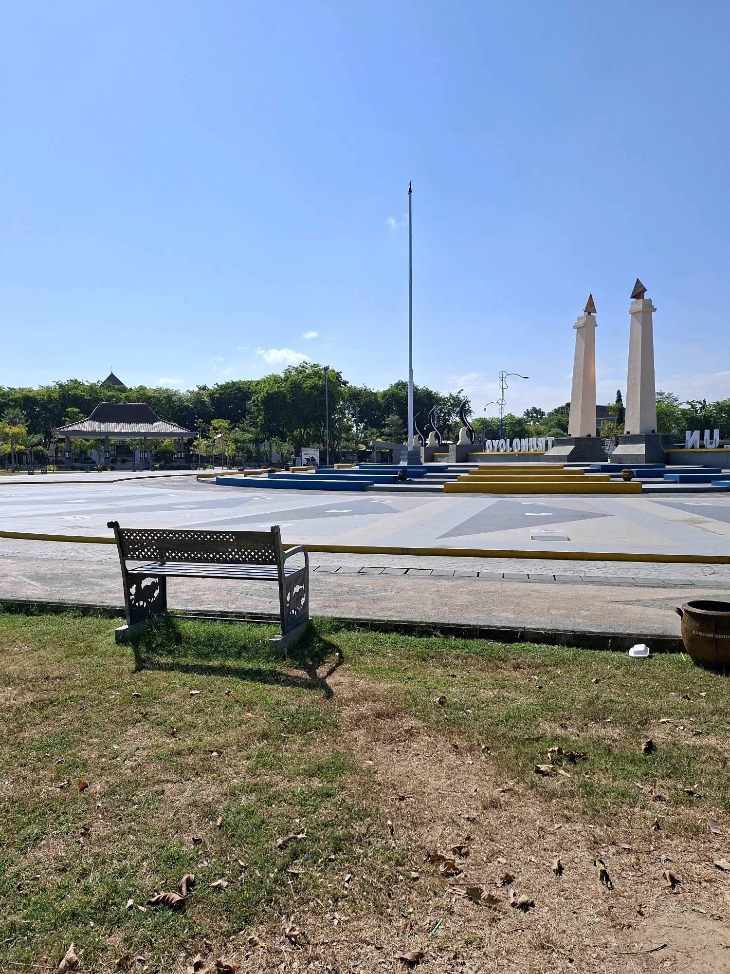 A tranquil outdoor scene unfolds under a clear blue sky, capturing a prominent monument and its surrounding public space in Gunung Sekar, Indonesia. In the foreground, a solitary, ornate metal bench with an elephant motif sits on a patch of parched, dry grass littered with fallen leaves. Beyond the bench, a vast, paved plaza features a striking architectural design with geometric patterns and bright yellow and blue accents. Dominating the mid-ground is the monument itself, characterized by two tall, cream-colored obelisks topped with conical shapes, likely representing significant landmarks or historical elements of the city. A flagpole stands tall to the left of the obelisks. Behind the monument, lush green trees form a verdant backdrop, with a traditional Indonesian-style pavilion visible through the foliage. The overall environment suggests a well-maintained public park or memorial site, bathed in bright sunlight, indicative of a warm, clear day. A brown ceramic trash receptacle labeled "SAMPAH KERING" (Dry Waste) is visible on the right side of the frame, with a discarded white styrofoam container nearby, suggesting recent human activity.