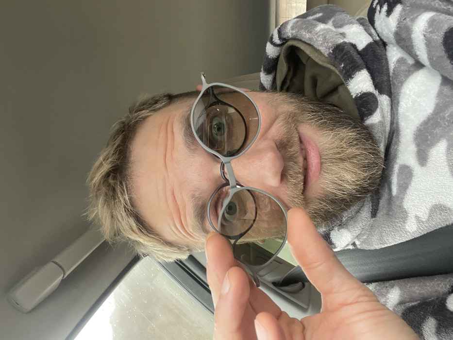 An indoor, frontal, mid-shot portrait captures a male subject situated within a vehicle in Bukovik, Montenegro. The subject exhibits fair skin, short light-colored hair, and a full beard and mustache. He is wearing round, light grey-framed eyeglasses. His left hand is raised, holding a small, rectangular, lightly tinted lens or magnetic attachment, which is positioned next to the left lens of his primary glasses. The subject's eyes are open wide, and his lips are slightly upturned in a smile, with some teeth partially visible on the right side of his mouth. He is dressed in a grey, white, and black camouflage-patterned fluffy hoodie, revealing an olive-green interior lining within the hood. The background depicts the light-colored interior headliner of the vehicle and a blurred exterior view through a window on the left, consistent with an overcast or rainy condition.