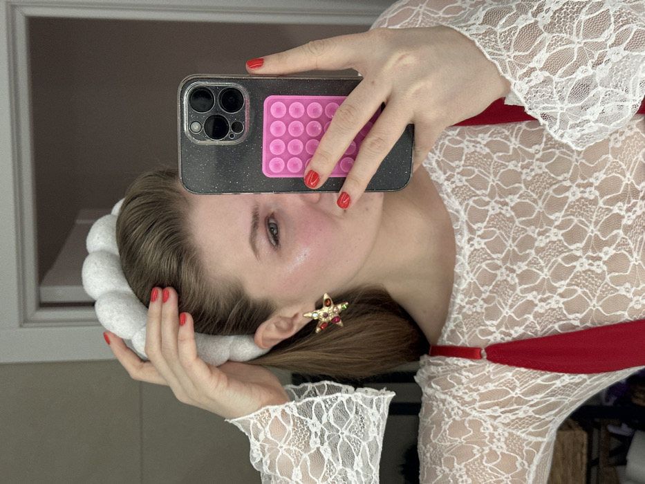 A woman takes a selfie in a mirror. She is fair-skinned with light brown hair pulled back, featuring a white padded headband. A star-shaped earring with red and green gems is visible. She wears a white lace top with red straps. Her nails are painted red. The phone is dark-colored with a pink bubble-pop grip attached. The background includes a window and indistinct furnishings.
