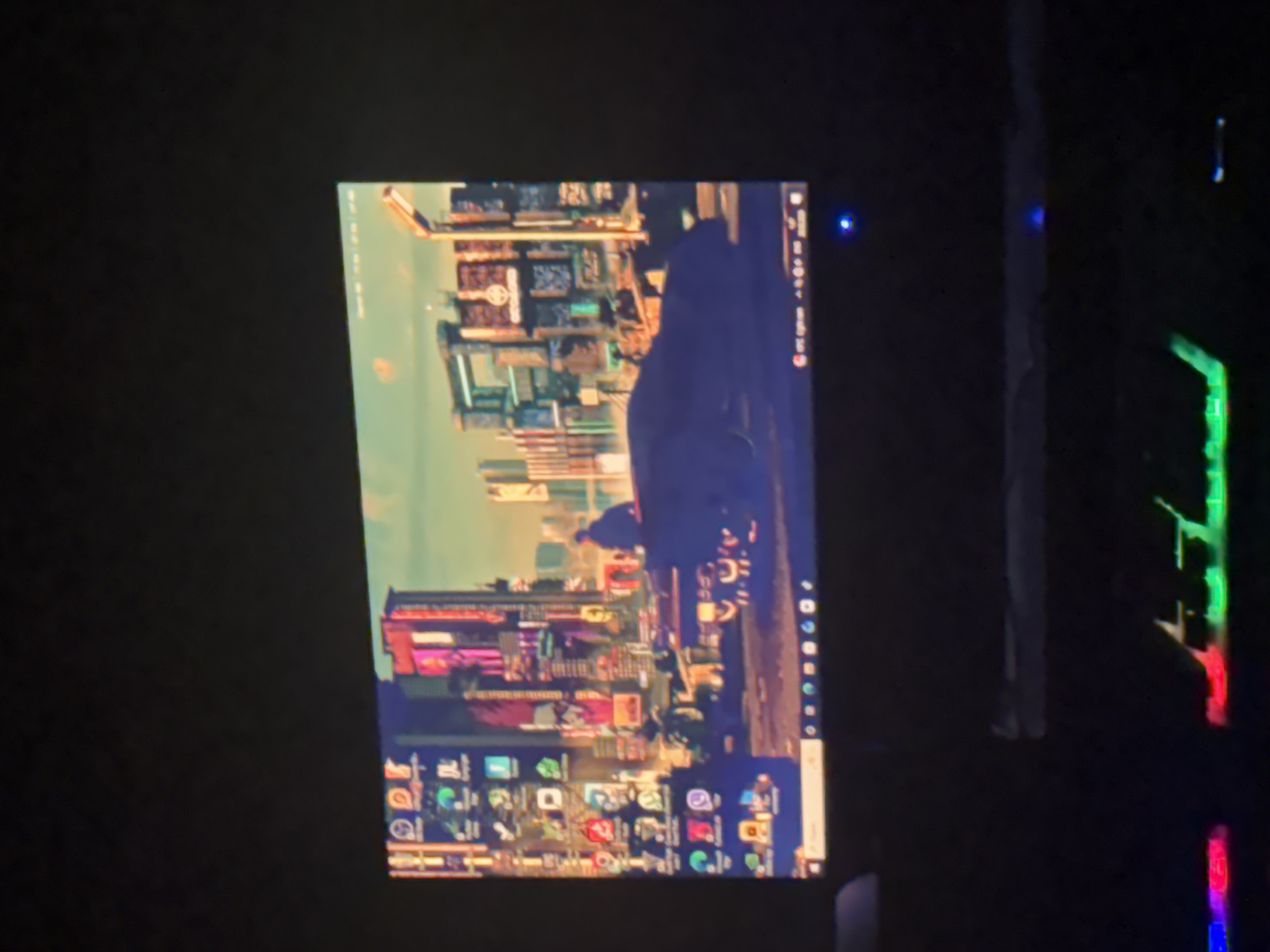 The image shows a computer monitor displaying a wallpaper with a city scene. The city has tall buildings and appears to be futuristic with neon lights and holographic advertisements. The monitor is surrounded by a dark environment. To the right of the monitor, there is a computer keyboard with multi-colored lights. I cannot identify this to be the city of Yerevan, Armenia, because there is no location information within the image.