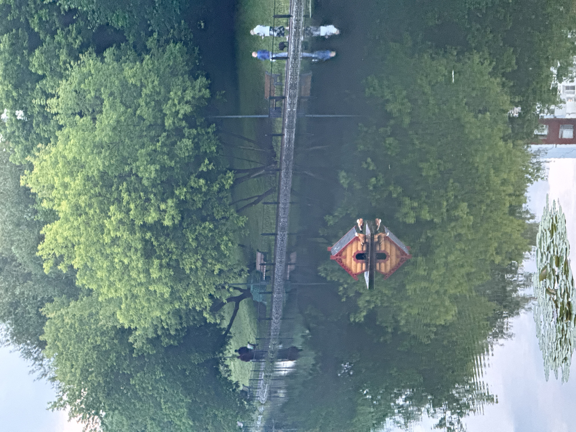 In a city park in Moscow, Russia, a small, red and yellow, wooden birdhouse with a peaked roof floats on a calm lake. The birdhouse is situated on a flat, dark platform and has a dark-colored opening. Two decorative duck figures are perched on the roof of the birdhouse. The lake's surface reflects the surrounding lush green trees and a few buildings. Along the shore, a black metal fence separates the water from a paved walkway. On the walkway, two people are walking a dog, and further down, two individuals stand by a bench. The ground beyond the walkway is covered in grass, and more trees dominate the background. Lily pads are visible in the foreground of the lake.