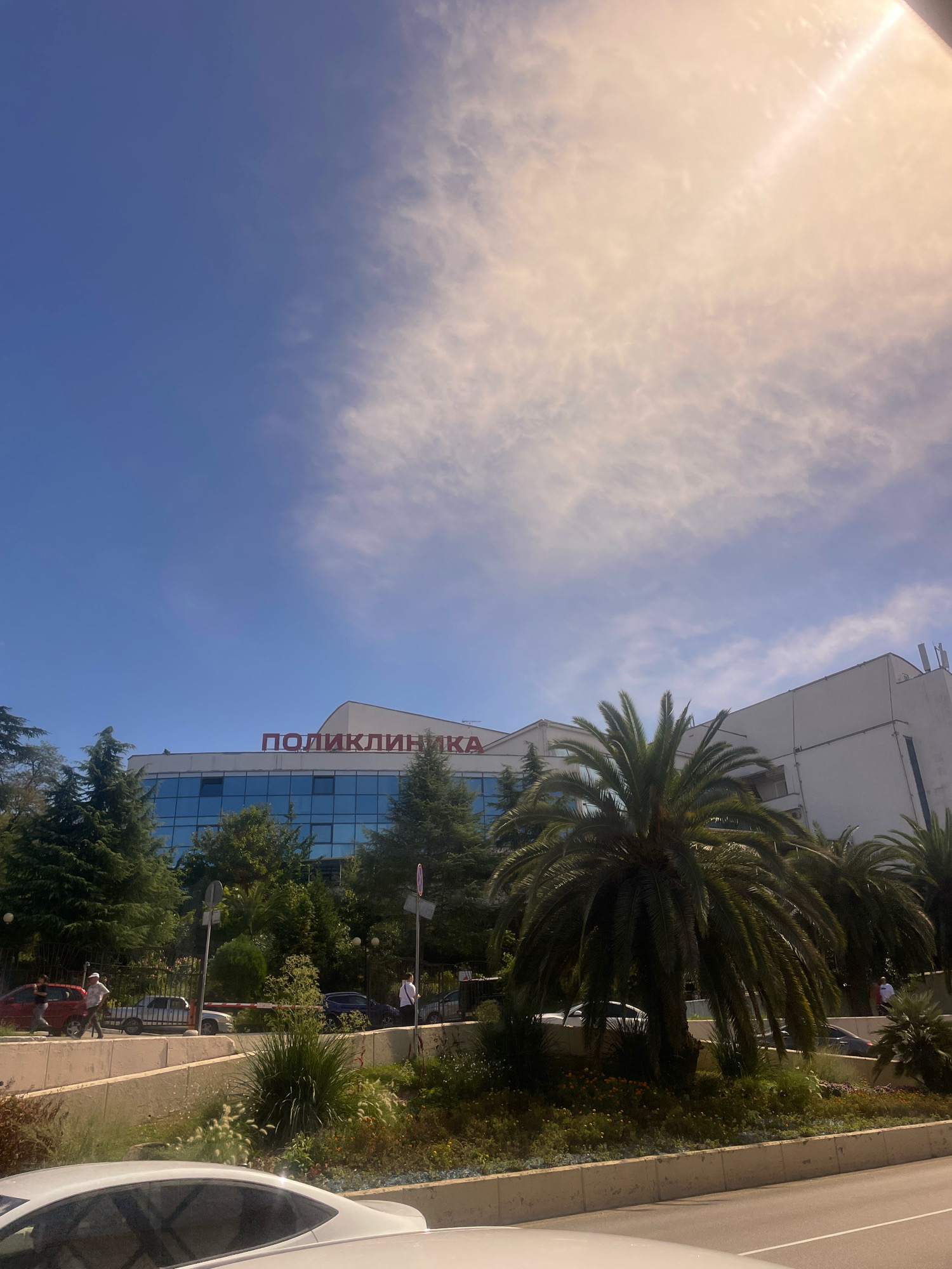The image shows a building with "ПОЛИКЛИНИКА" (Polyclinic) written in red letters on its facade. The building appears to be a medical facility. It is surrounded by lush greenery, including palm trees and coniferous trees, suggesting a warm climate. The sky is blue with some scattered clouds, and the sun is shining, casting a bright glare in the upper right portion of the image. In the foreground, a road is visible with several cars parked or driving by. People are seen walking near the building. The overall scene suggests a sunny day in a city with a pleasant environment. This building is located in Sochi, Russia.