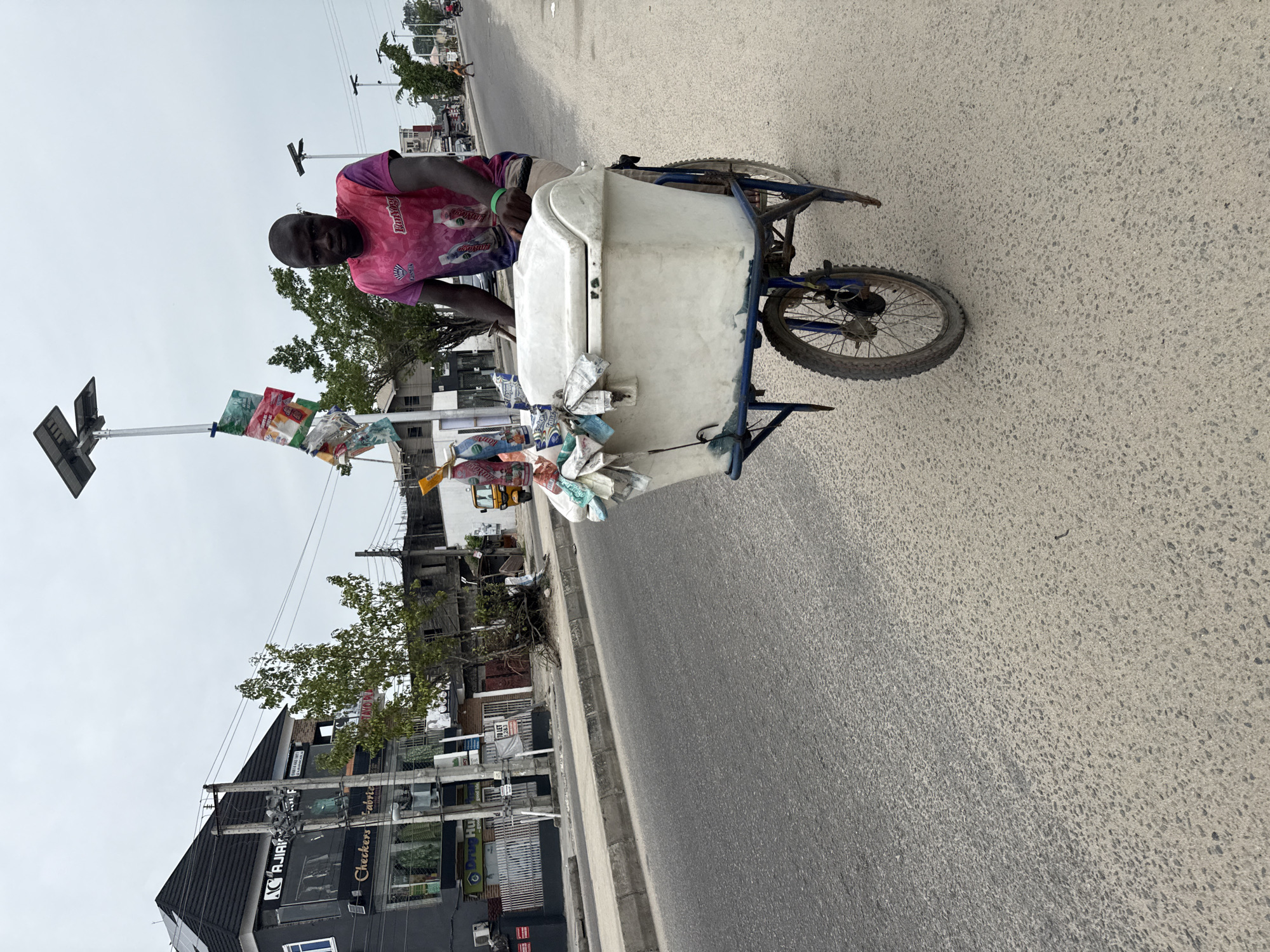 An African man sits on a three-wheeled cart, which he appears to be using to sell packaged goods. The cart is topped by a large, white container with blue trim, on which packages are tied to the front. Additional packages are attached to a vertical pole affixed to the cart. The road is paved and there are urban buildings in the background. The scene appears to be Ado, Nigeria, based on storefront signage in the background.