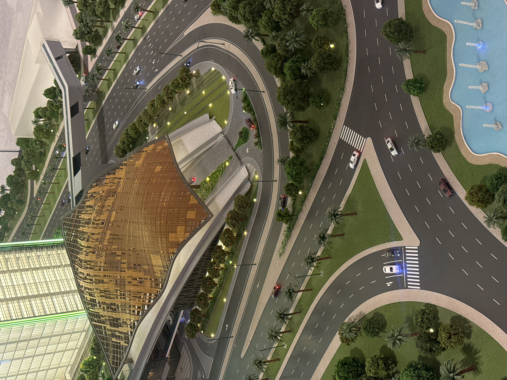 This is an aerial perspective photograph of a scale model, likely an architectural or city planning model. The subject matter includes miniature buildings, roads, landscaping, and vehicles.
A significant building with a gold-colored, curvilinear roof is centrally located in the frame. Nearby, another modern building has a gridded roof. Roads surround the structures, featuring miniature cars, trees, streetlights, and pedestrian walkways.
On the right side, a body of water with blue fountain displays is visible. The overall environment is well-manicured, with lush greenery and palm trees.
This appears to be a scale representation of a modern urban area, potentially located in Dubai, United Arab Emirates, given the architectural style, landscaping, and urban planning elements present in the model.