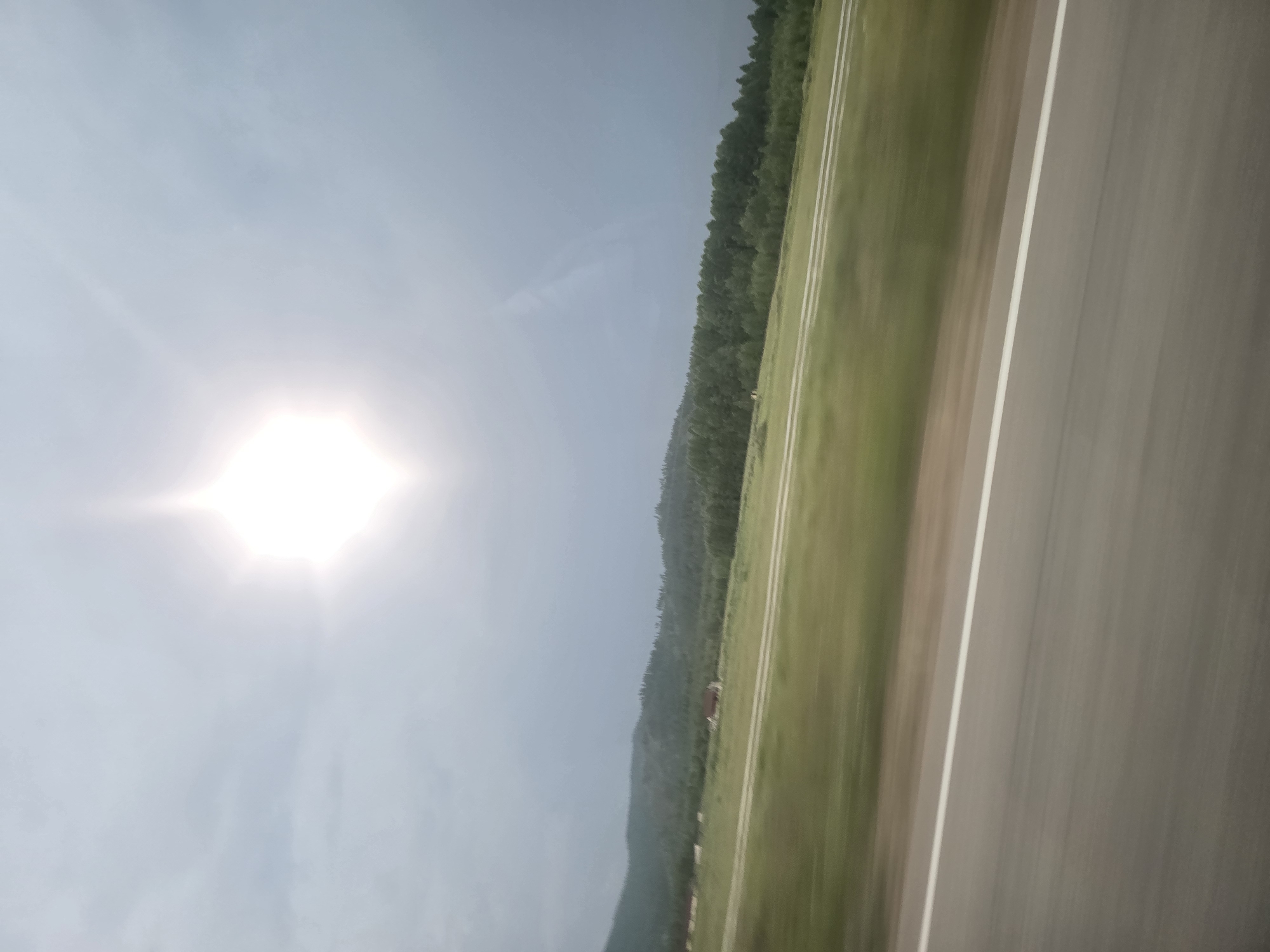 The image depicts a view from a moving vehicle, likely a car, looking out over a landscape. The foreground is dominated by a blurred asphalt road with a white lane marker. Beyond the road is a field of green grass, also appearing blurred due to motion. Further in the background, there is a dense line of trees, likely a forest, and rolling hills or mountains. The sky is overcast with a bright, star-shaped sun peeking through the clouds, creating a lens flare effect. The scene appears to be rural, consistent with the outskirts of a settlement like Angyr, Russia, characterized by open spaces, vegetation, and a road network. No people are visible in the image.