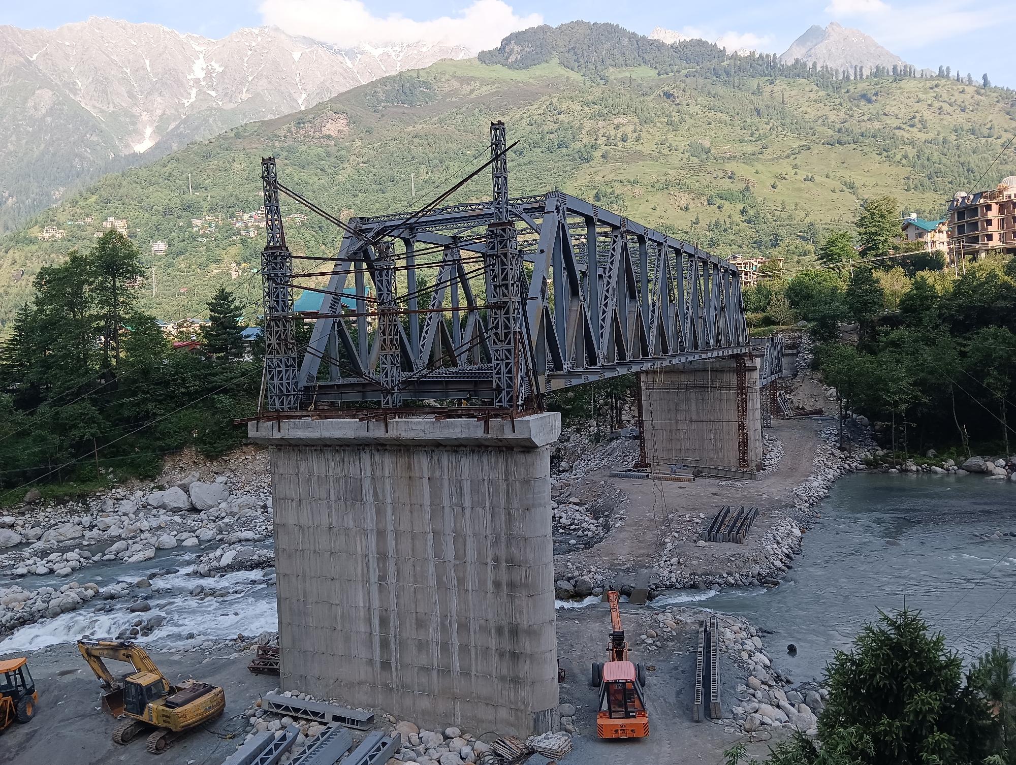 The image captures a construction site for a bridge over a river in Kulang, India. In the foreground, a large concrete pillar supports a grey steel truss bridge, which appears to be under construction. Yellow and orange construction vehicles, an excavator and a crane, are parked on the riverbank. The river flows with clear water over a rocky bed. The background features lush green mountains with scattered buildings and snow-capped peaks in the distance, indicating a mountainous region. The sky is partly cloudy. The scene depicts an active construction environment against a natural, scenic backdrop.
