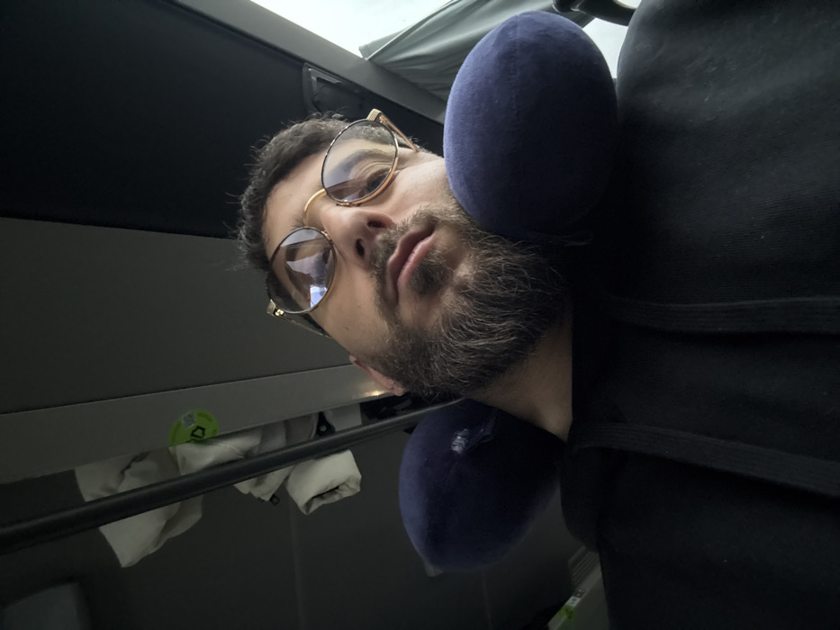 A low-angle, close-up photograph captures an adult male resting inside a public transportation vehicle, likely a bus or train, in Rosny-sur-Seine, France. The subject, positioned centrally, has a short dark beard with grey highlights and short dark hair. He wears gold-framed glasses with a prominent upper brow line, reflecting light. His eyes appear heavy-lidded or closed, suggesting sleep or rest. A dark blue, U-shaped travel neck pillow supports his head. He is dressed in a black hooded sweatshirt with visible drawstrings. In the upper left background, an overhead storage compartment contains several folded white fabric items and a small green circular sticker with a white logo. A muted, diffused light source is visible through a window on the upper right, indicative of an overcast sky or interior lighting.