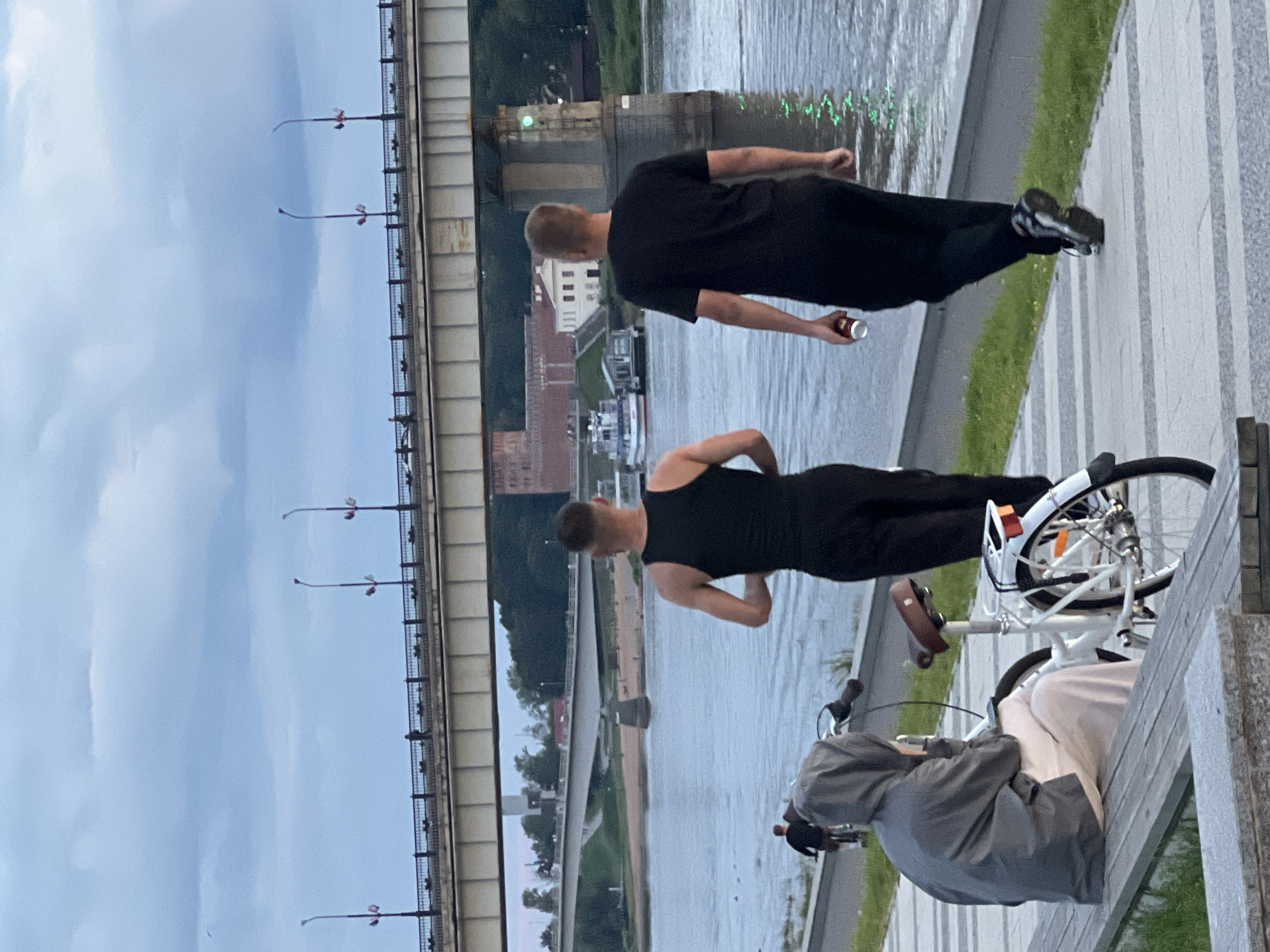 The image depicts a riverside scene in Veliky Novgorod, Russia, likely near the Yaroslav's Court and Kremlin complex, given the prominent bridge and red-brick structures visible in the background. In the foreground, three individuals are present. On the left, a person wearing a grey hooded jacket and light-colored pants sits on a wooden bench next to a white bicycle. To the right of the bench, two men are walking away from the camera along a paved pathway. The man in the center wears a black tank top and black pants and is in motion, appearing to be jogging or walking briskly. He is holding a red and white can in his right hand. The man to his right wears a black t-shirt and black pants and is also walking away. The pathway runs alongside a wide river, where several boats are docked or moored. A large bridge spans the river in the middle ground, with lampposts lining its railing. The sky is overcast with dramatic clouds. In the distance, red-brick buildings and green trees are visible on the riverbank. A smaller bridge is also visible further upstream. A person on a bicycle is seen in the far left of the image.