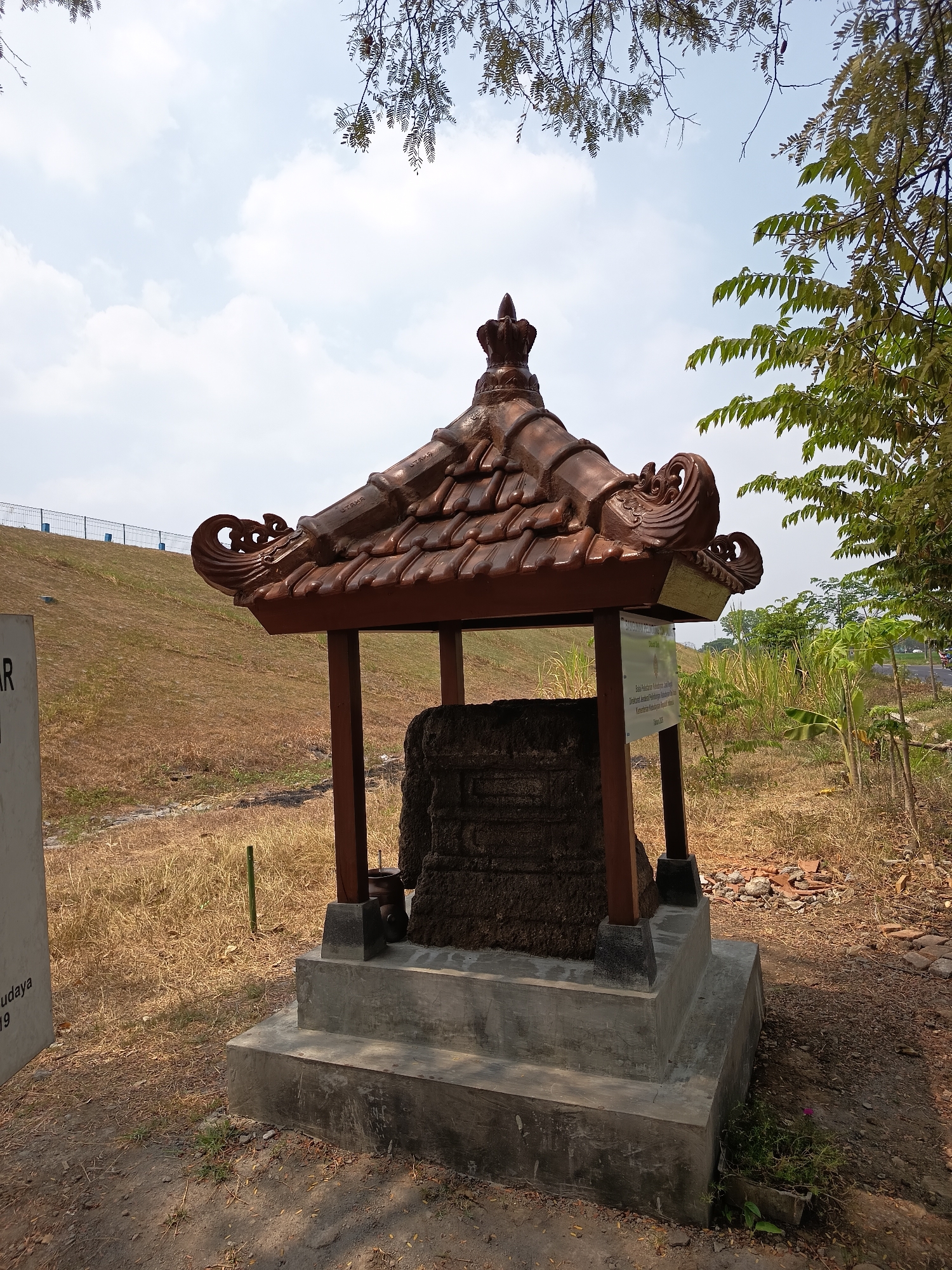 In the city of Lalung, Indonesia, an outdoor scene features a stone monument under a decorative tiled roof. The monument itself appears to be a large, weathered stone with visible inscriptions or carvings. It is housed within a simple wooden structure with a concrete base and steps. A white signpost stands to the left of the monument, and a sign is also attached to one of the wooden posts of the structure, providing information about the monument. The environment surrounding the monument is dry and grassy, with some foliage and trees visible in the background. A road and a body of water with a fence are also visible in the distance, suggesting a somewhat developed area. The sky is partly cloudy.
