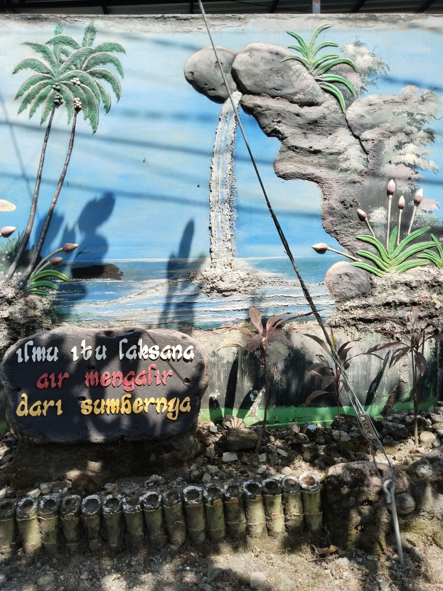 This image shows a three-dimensional mural, possibly in Sepat, Indonesia. The mural depicts a landscape scene with a waterfall, rocks, stylized plants, and a painted backdrop suggesting the sky and water. In the lower foreground, there is a plaque with an inscription, possibly in Indonesian, with white, red, and yellow lettering. The base of the mural is decorated with vertical pieces of bamboo or similar material. A metal cable runs diagonally across the scene. Shadows are cast across the image. The scene is outdoors, and appears to be on the side of a building or retaining wall.