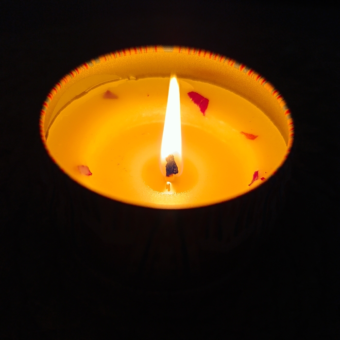 The image presents a close-up, high-angle view of a lit candle. The candle is contained within a round, metallic vessel, the rim of which displays faint, multicolored striations. The candle wax is a solid, opaque yellow-orange, with a molten pool surrounding a dark, upright wick at its center. A single, bright, teardrop-shaped flame, primarily yellow with a white core, burns steadily from the wick. Scattered across the surface of the melted wax and partially embedded in the solid wax are several small, irregular dark red fragments, consistent with dried flower petals. The background is uniformly black, indicating a dark environment where the candle flame is the sole source of illumination. No individuals are visible, and the image contains no visual information to identify a specific location in the city of Марковский, Russia.