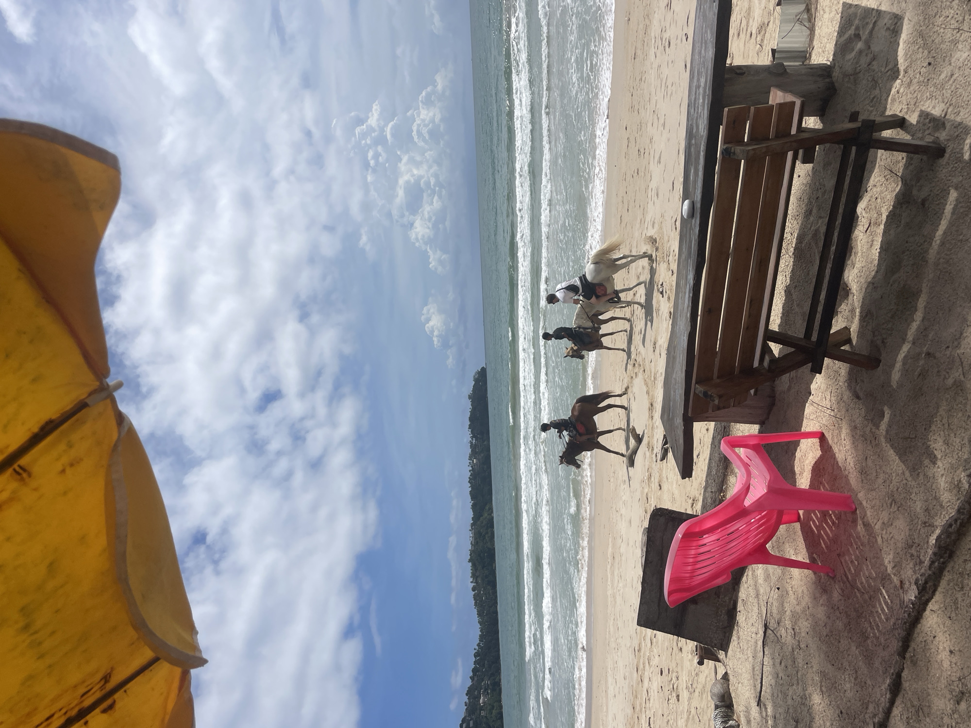 The image shows a beach scene near Choeng Thale, Thailand. Three people are riding horses on the beach close to the water. A yellow umbrella is visible in the foreground on the left. A wooden bench and a red plastic chair are situated on the right side of the frame. The sky is blue with scattered clouds. The water is relatively calm. The land is visible in the distance on the opposite side of the water.