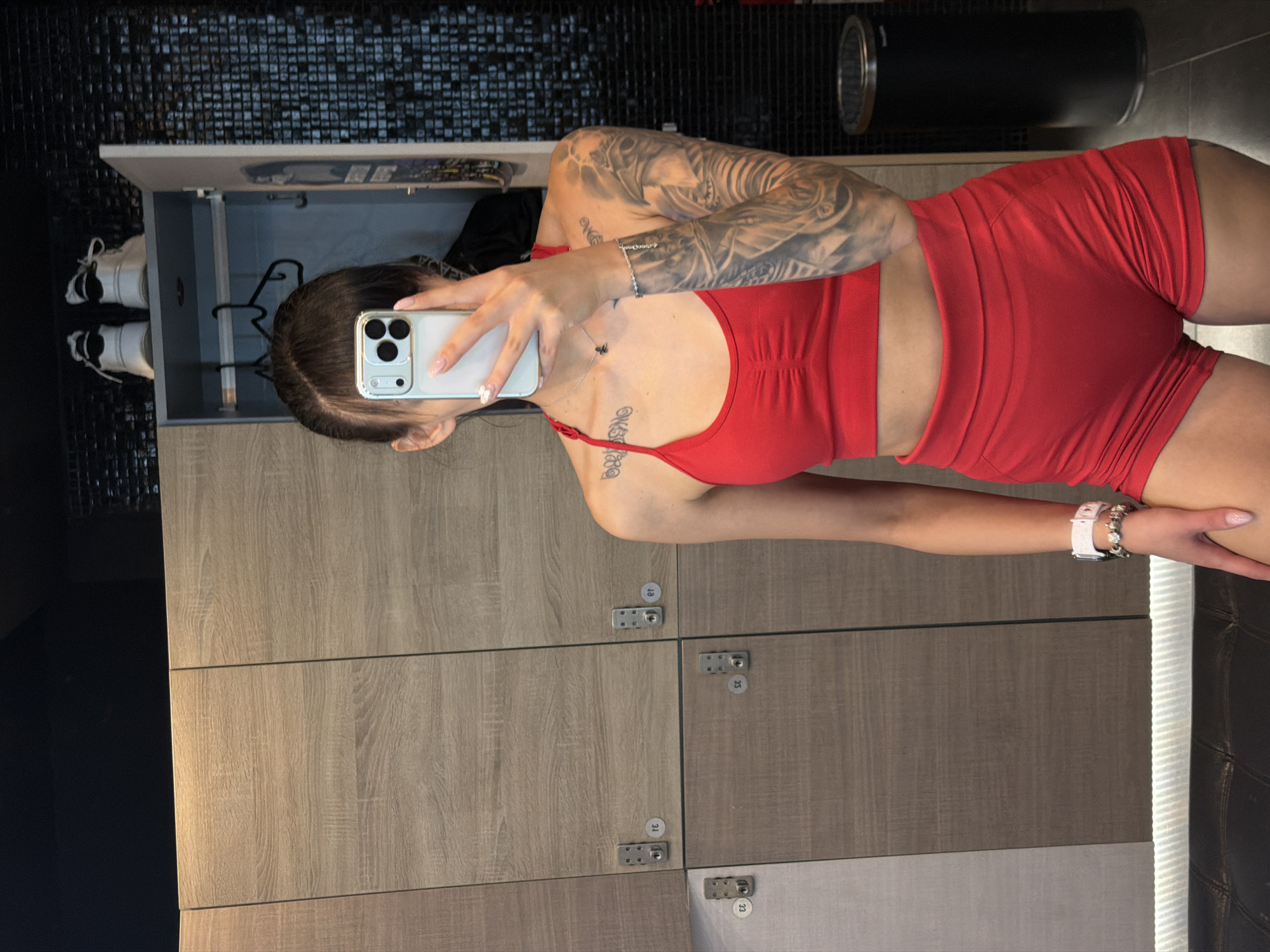 A young adult female stands in a locker room, taking a mirror selfie with her phone. She is wearing a red sports bra and matching red shorts. Her left arm, visible in the frame, is heavily tattooed with an elephant design. She has a necklace and a bracelet on her left wrist, and a watch on her right. The background consists of wooden lockers. In the upper right corner, a black bin is visible. The overall environment suggests a gym or a similar facility.