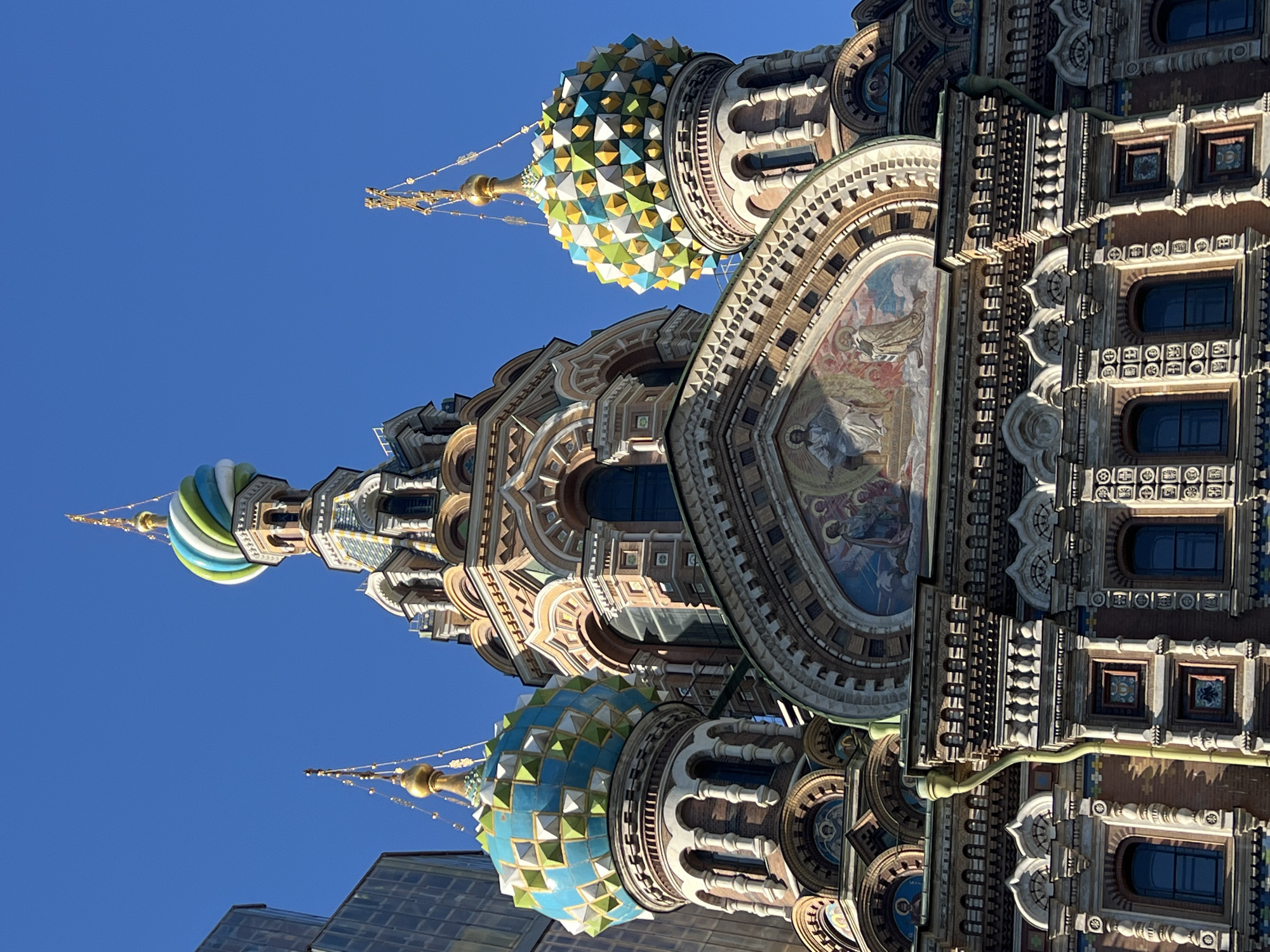 The image shows a close-up, upward-angled view of the Church of the Savior on Spilled Blood in Saint Petersburg, Russia, against a clear blue sky. The elaborate architectural details are prominently displayed, including the onion-shaped domes with their colorful, geometric patterns. The building's facade features mosaics, ornate windows, and intricate masonry. A modern building is visible at the bottom left corner. The photo does not depict any people.