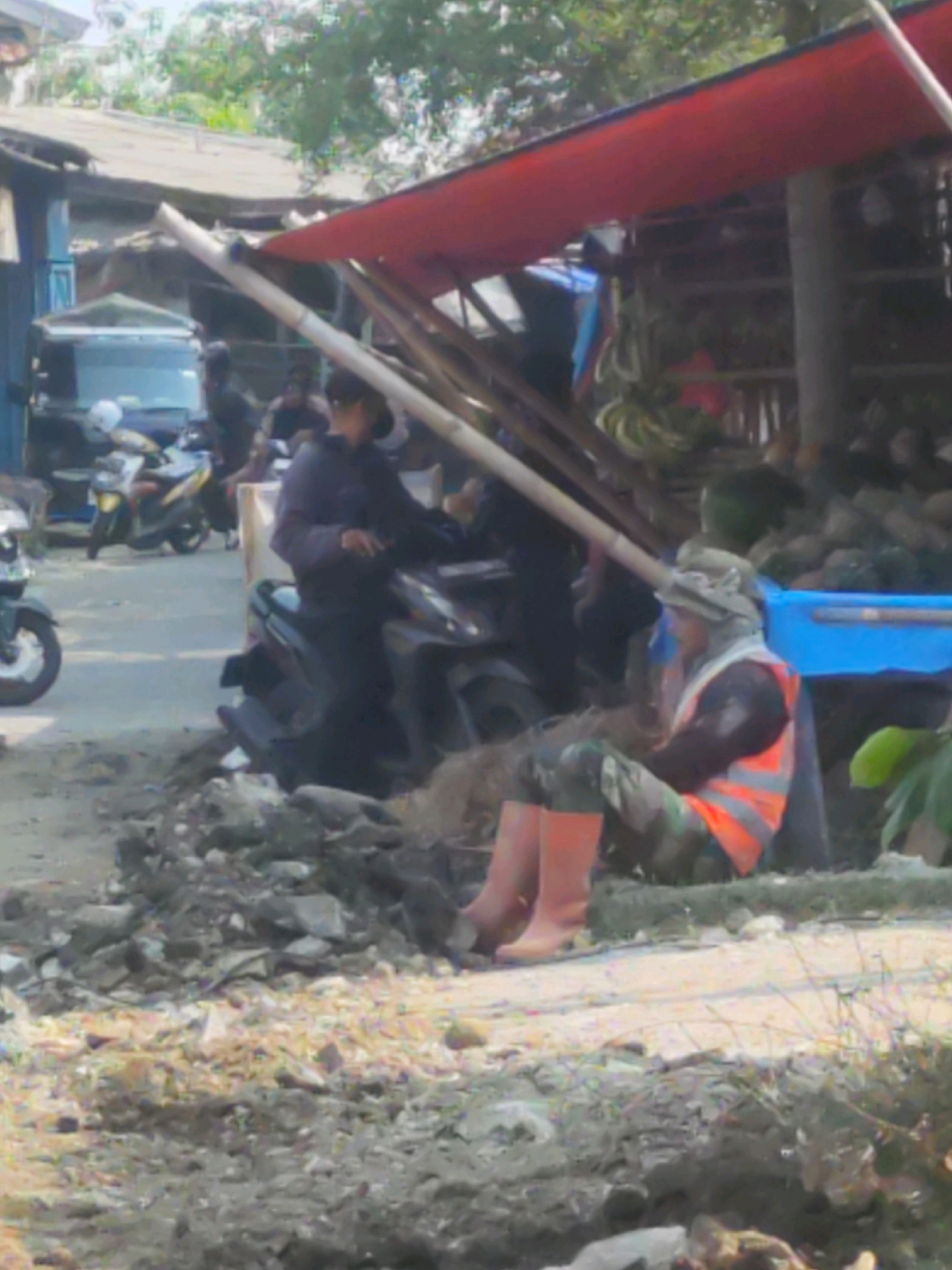 A medium-shot, low-angle, slightly blurry photograph depicts an outdoor market scene in Karawang, Indonesia. In the foreground, a person wearing a camouflage-patterned long-sleeved shirt, an orange safety vest, and orange rubber boots is sitting on the ground. The person has a hat covering their head and is looking downwards. Behind them, several motorcycles are parked. A red awning, supported by bamboo poles, extends over a stall area filled with what appear to be bananas and other produce. The ground is uneven and covered with dirt, rocks, and debris. In the background, buildings with corrugated metal roofs and trees are visible. The overall impression is one of a bustling, informal marketplace.