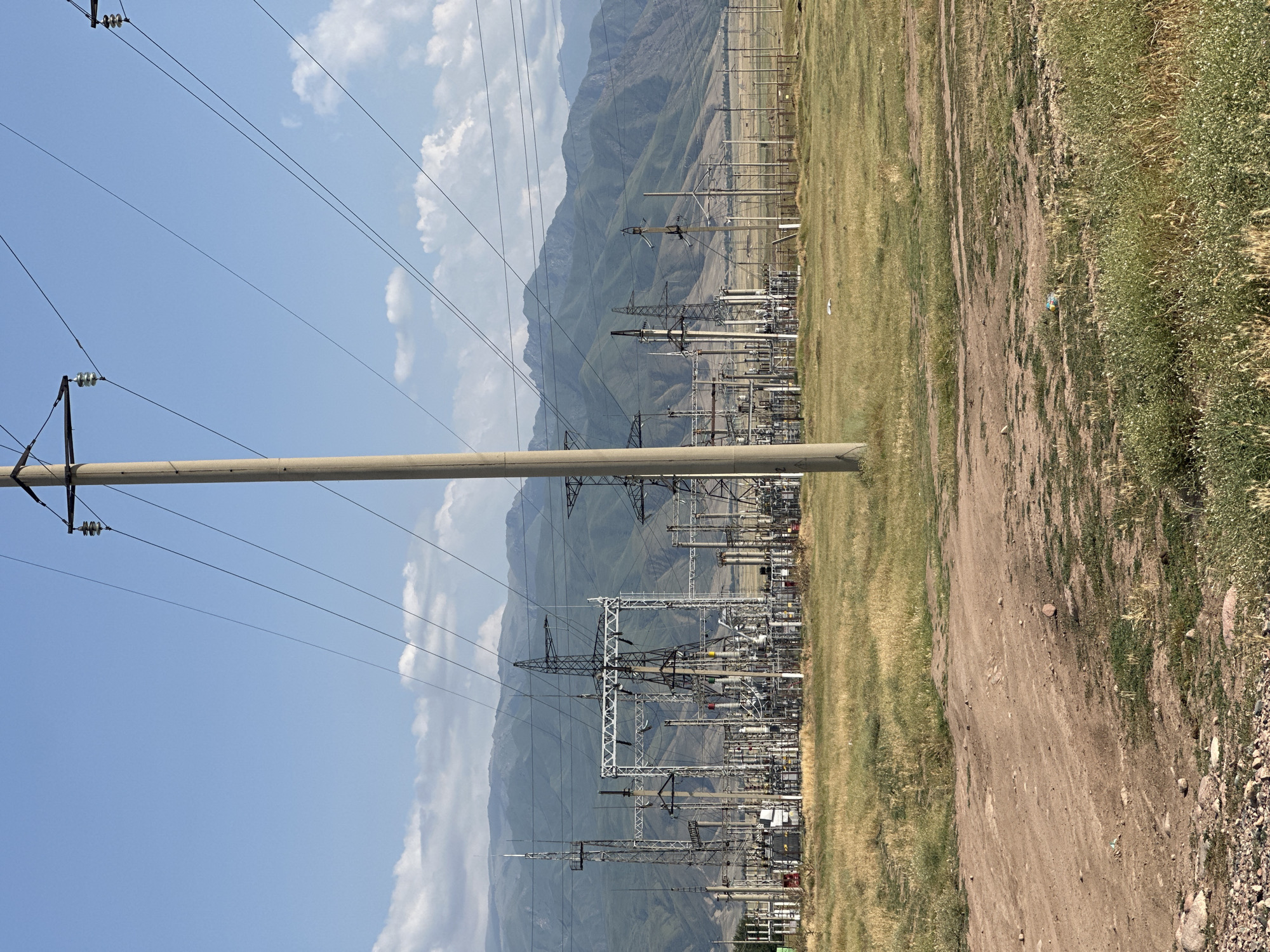 This media file depicts a power substation set against a backdrop of mountainous terrain and a clear blue sky with scattered clouds. The substation is a complex arrangement of towers, transformers, and electrical lines, suggesting an active electrical infrastructure. A large concrete pole and numerous power lines dominate the foreground, extending across the frame and connecting to the substation in the midground.
The scene is set in a rural or semi-rural area, indicated by the presence of dry, grassy fields and a dirt path running alongside the substation. The vegetation appears sun-baked, and the overall lighting suggests it is daytime, likely a sunny afternoon. The distant mountains are layered and appear green and brown, hinting at the geographical location in Kazakhstan, specifically within the Sarymoldayev rural district.
There are no people visible in the frame, and the overall mood is one of industrial infrastructure in a natural landscape. The focus is on the electrical equipment and its integration into the environment. The image is taken from a slightly low angle, emphasizing the height of the power lines and the scale of the substation. No visible text is present in the image.
