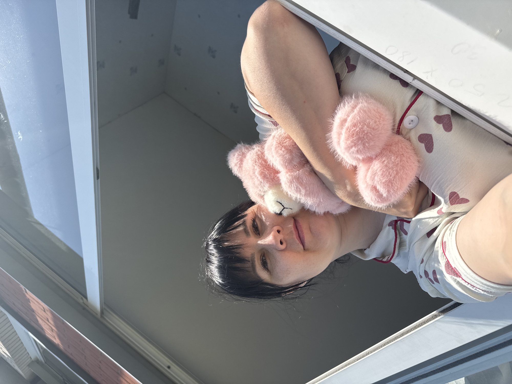 A woman is holding a pink stuffed animal close to her face while looking down into the camera from an open window. She is wearing pajamas patterned with hearts. The window frame is white and the background outside the window is bright. It is not possible to determine the location in Zav'yalovo, Russia, based on the image.