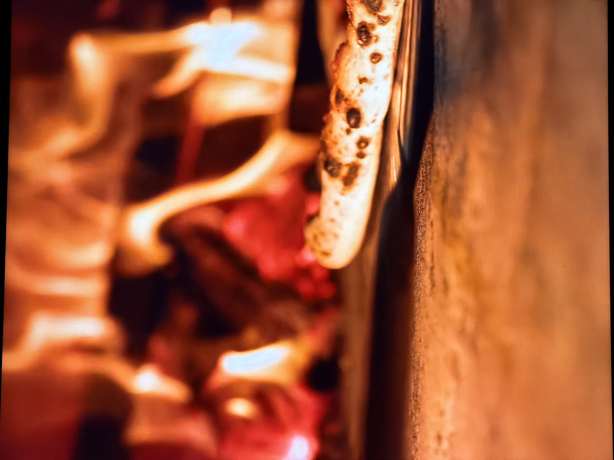 The image displays a close-up view of a pizza being cooked in a wood-fired oven. Flames and glowing embers are visible in the background, creating an orange and yellow ambiance. A pizza peel is holding a pizza, which has a crust dotted with dark spots, indicating it is being cooked at high heat. The surface of the oven, likely made of stone or brick, is in the foreground, showing a rough texture. The image does not contain any identifiable landmarks or elements that would allow for a specific location in Chirchik, Uzbekistan to be determined.