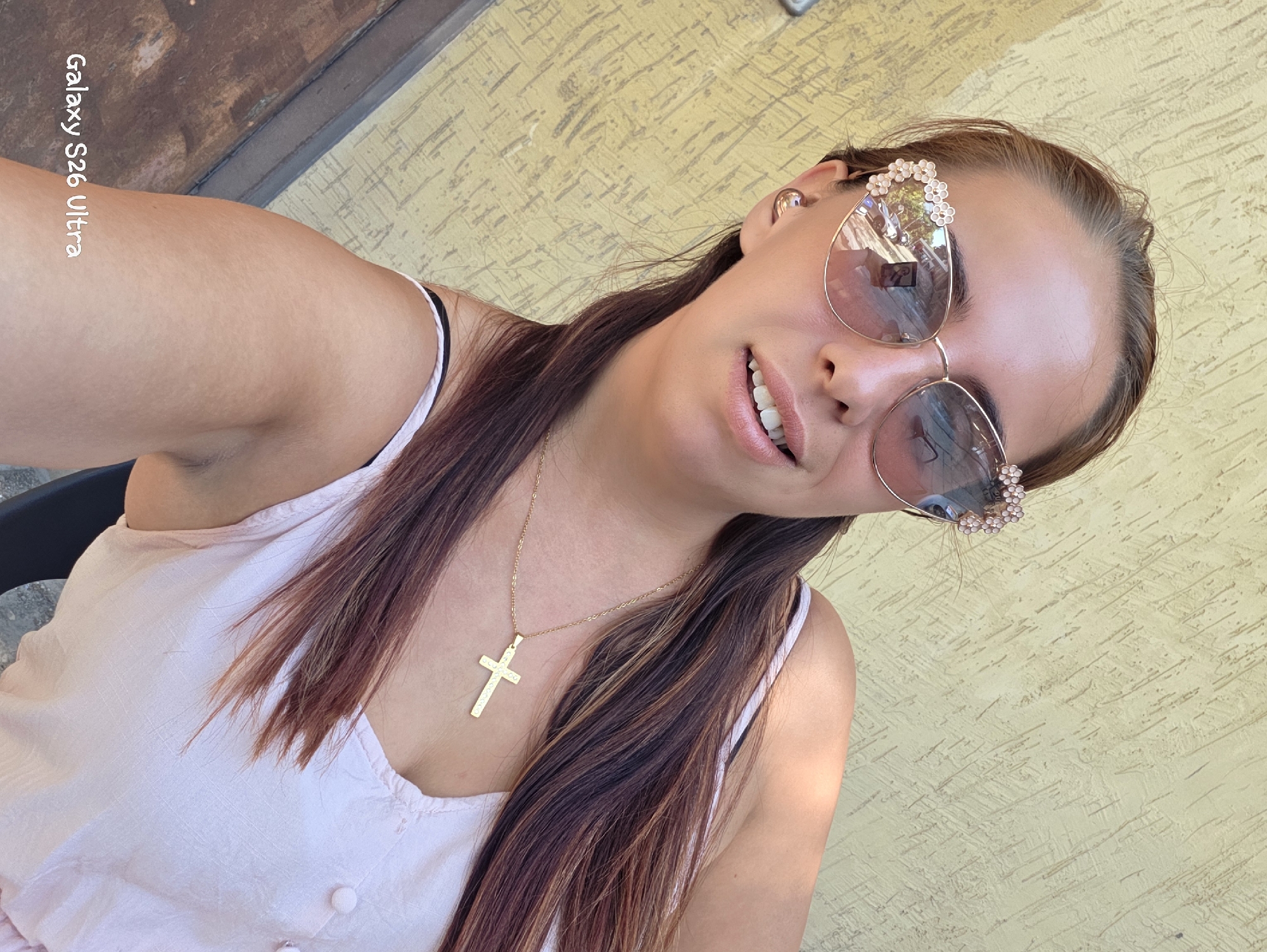 A close-up selfie shows a woman wearing large, ornate sunglasses with floral accents and a gold cross necklace. She is outdoors, with a textured yellow wall in the background. The "Galaxy S26 Ultra" watermark is visible in the bottom left corner.