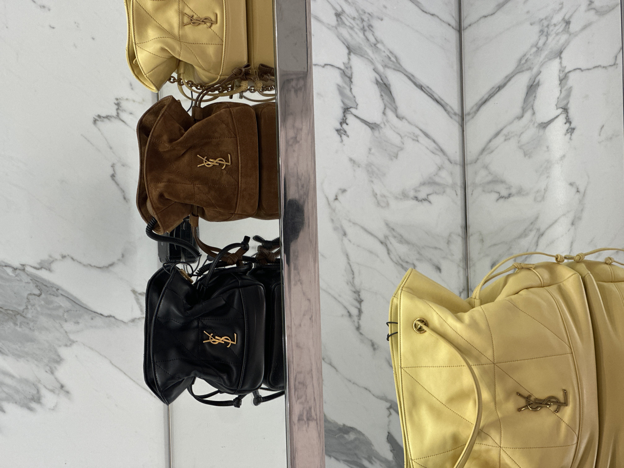 The image displays a luxury retail setting, presumed to be within a Yves Saint Laurent (YSL) boutique or a high-end department store in London, United Kingdom. The environment features walls clad in white marble with prominent grey veining. Two reflective metallic shelves are visible. On the upper shelf, three YSL drawstring bucket bags are presented side-by-side. From left to right, these bags are black quilted leather, brown quilted suede, and pale yellow quilted leather, each adorned with a gold YSL monogram logo on the front. The pale yellow bag is partially cropped on the right. Below, on a lower, also reflective, shelf, a portion of a larger pale yellow quilted leather YSL tote bag with a gold monogram is visible in the bottom left quadrant of the frame. No individuals are present, and all items are static display pieces.