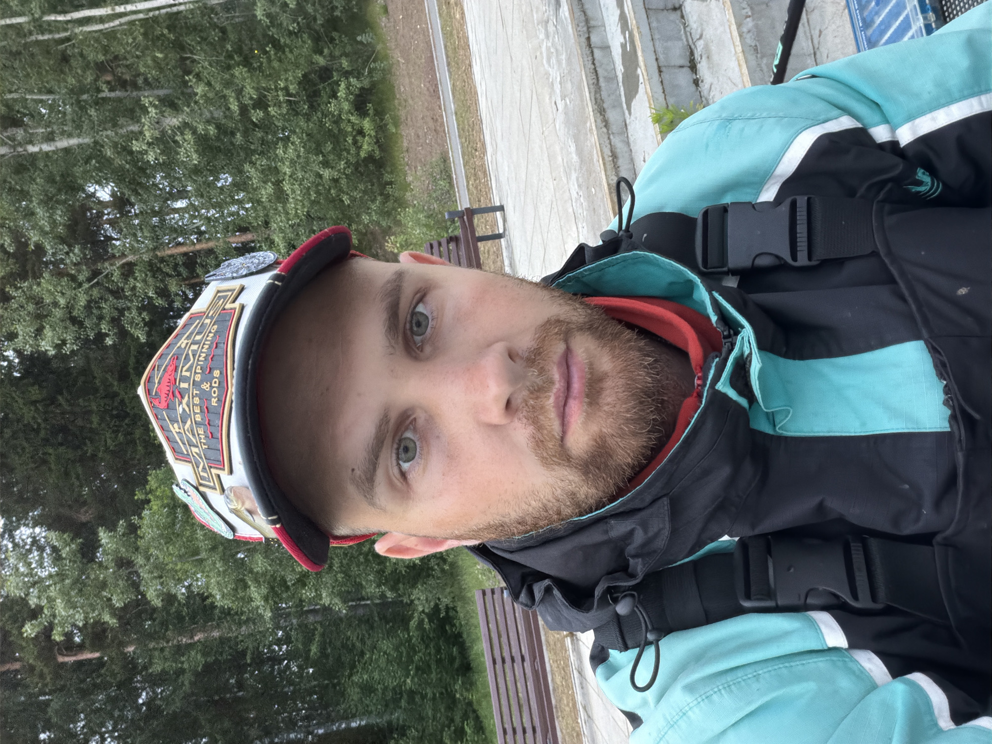 A male subject, estimated to be in his late 20s to early 30s, is depicted in a close-up, chest-up outdoor portrait, facing directly towards the camera. The subject has a fair complexion, light greenish-gray eyes, a light brown to reddish-brown full beard and mustache, and matching eyebrows. He wears a baseball cap with a red brim, a white front panel, and a black mesh rear. The front of the cap displays an embroidered design that reads "MAXIMUS THE BEST SPINNING & RODS" in red and black lettering, flanked by a red lobster/crawfish emblem on the left and a fish lure on the right. Additional decorative pins, including a green lizard or snake and a circular silver emblem, are affixed to the cap. The subject is wearing a two-tone outdoor jacket, predominantly light teal/aqua blue on the upper and outer sections, with significant black panelling on the chest and arms. A red inner collar is visible. A black strap with a black plastic quick-release buckle is prominent over his left shoulder, indicating a harness or part of a vest/waders system.
The background is an outdoor environment in what is identified as Chaikovsky, Russia. Dense green foliage and trees, including both conifers and deciduous varieties, form the backdrop. Two dark brown slatted wooden benches with dark frames are positioned on either side of the subject in the mid-ground. The ground in the foreground consists of light-colored paved sections, possibly flagstones or concrete, leading to a series of light-colored steps on the lower right, which appear damp in areas.
