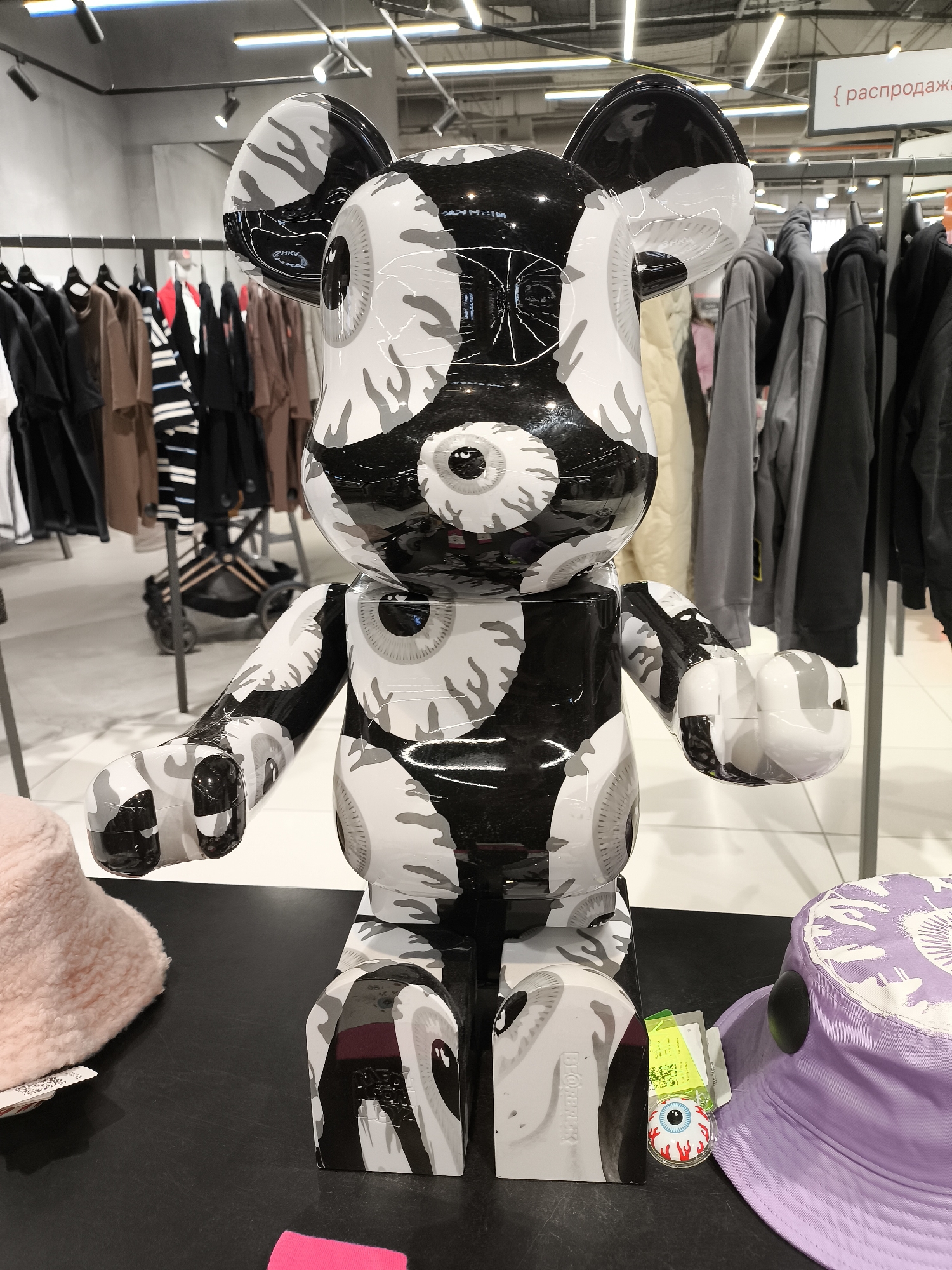 The image shows a large, glossy black and white Bearbrick figure with an "eye" design standing on a black surface. The figure is in a retail environment, surrounded by racks of clothing in the background. A light pink furry hat is to the left of the figure, and a light purple bucket hat is to the right. A small, cartoon-like eyeball image is visible at the base of the figure near the bucket hat. The location is a store in an unknown city.