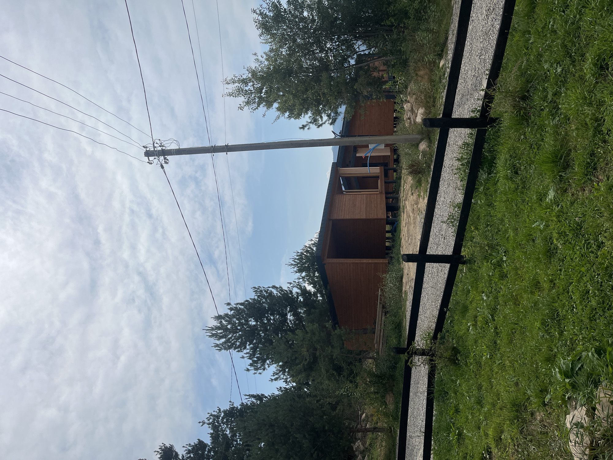 This is a wide shot of a rural scene near Mykulychyn, Ukraine, featuring a modern wooden house or cabin. The house is situated amidst pine trees and lush green grass. A dark wooden fence runs horizontally across the foreground, separating a gravel path from the grassy area. Power lines stretch across the cloudy sky, supported by a wooden pole. The house appears to be unfinished or under construction, with exposed wooden beams and open sections. One of the cabins has a blue pipe extending from its wall. The overall impression is one of a serene, natural setting with a touch of modern architecture.