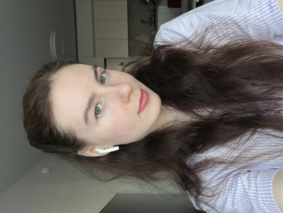 A female individual is depicted from the chest up in an indoor residential setting. She has fair skin, blue-green eyes, and dark brown wavy hair, partially pulled back from her face, with strands falling over her shoulders. She wears pinkish-red lipstick and a light purple and white striped shirt adorned with small purple bow patterns. A white Apple AirPods earbud is visible in her right ear. Her gaze is directed towards the camera. The background partially shows a modern kitchen area on the right, featuring white upper cabinets, a sink with a faucet, and a light-colored wall. A round, flush-mount light fixture is visible on the white ceiling. A red object is partially visible on a counter in the background. The specific location within the city of Ковалёво, Russia, is not discernible from the image content.