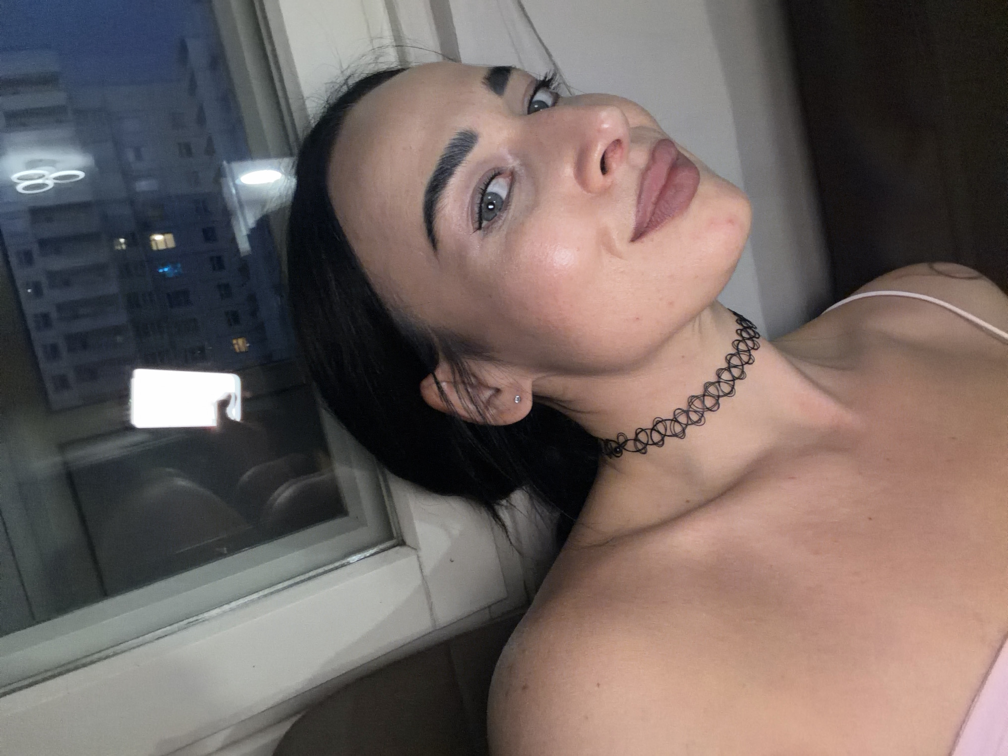 An angled, close-up portrait features a young woman with dark hair and striking green eyes, gazing directly at the viewer with a calm, confident expression. Her face is well-lit, highlighting her features, including prominent eyebrows and makeup, with a muted, rosy lip color. She wears a black, patterned choker necklace and a small stud earring in her visible ear. Her attire includes a light-colored, possibly pink, strap top, revealing her bare shoulder and collarbone.
The woman is situated indoors, positioned near a window that fills the left side of the frame. Outside the window, the scene is dark, indicating nighttime. A multi-story building is visible in the distance, with several windows illuminated, suggesting an urban or suburban setting. The window glass reflects various elements from the room, including a bright rectangular light source, possibly a mobile phone, and another bright, circular reflection. The window itself has a white frame.
The image captures a posed moment, likely a self-portrait or a close-up portrait. The setting appears to be a residence in Zanevka, Russia, during the late evening or night. There is no visible text in the image.
