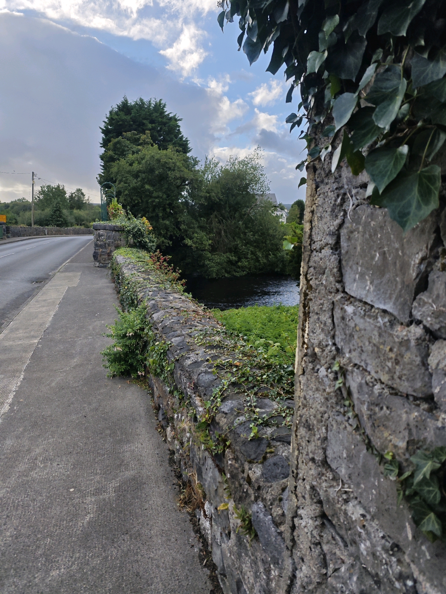 The image depicts a view from a pathway next to a stone wall bordering a body of water. A road curves away to the left, flanked by green foliage and trees. The sky above is partly cloudy with blue patches. The stone wall, covered in ivy and other vegetation, extends into the distance. A large tree stands prominently on the far side of the water, with more trees and a building visible beyond. The scene appears to be a picturesque spot along a road, possibly a bridge, in Oughterard, Ireland.