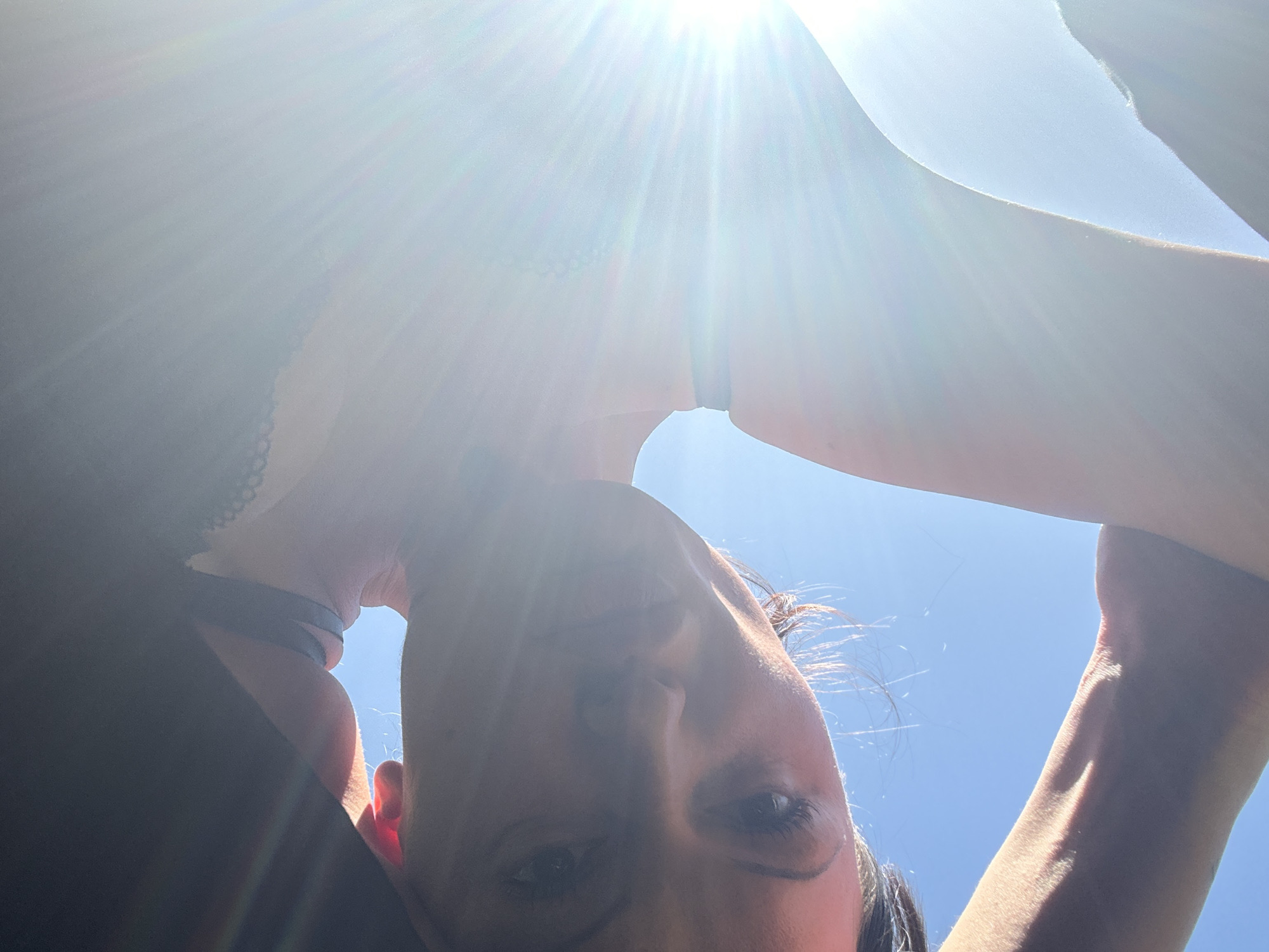 An upward-facing, low-angle selfie captures a person, likely a woman, looking directly into the camera from an inverted perspective. Her face is at the bottom of the frame, tilted slightly, with her dark hair visible on the sides and top of her head. Her eyes are open and directed towards the viewer.
The image is heavily backlit by an extremely bright, overhead sun, centered in the upper half of the frame. This intense light creates prominent lens flare and sun rays, obscuring much of the upper body and casting the face and arms largely in shadow, though highlights catch the edges of her features and arms. The sky behind her is a clear, bright blue, indicating good weather.
Her arms are raised, framing her head, and appear to be holding the camera above her. A dark top, possibly a bra or camisole, with a subtle lace detail is visible on her left side. A dark wristband is worn on her left wrist. The strong light emphasizes the texture of her skin on her arms.
The scene suggests an outdoor setting during daytime, likely midday, under a sunny sky. The overall impression is bright and high-contrast due to the direct sunlight. The location context provided is Tunbridge Wells, United Kingdom. No visible text is present in the image.