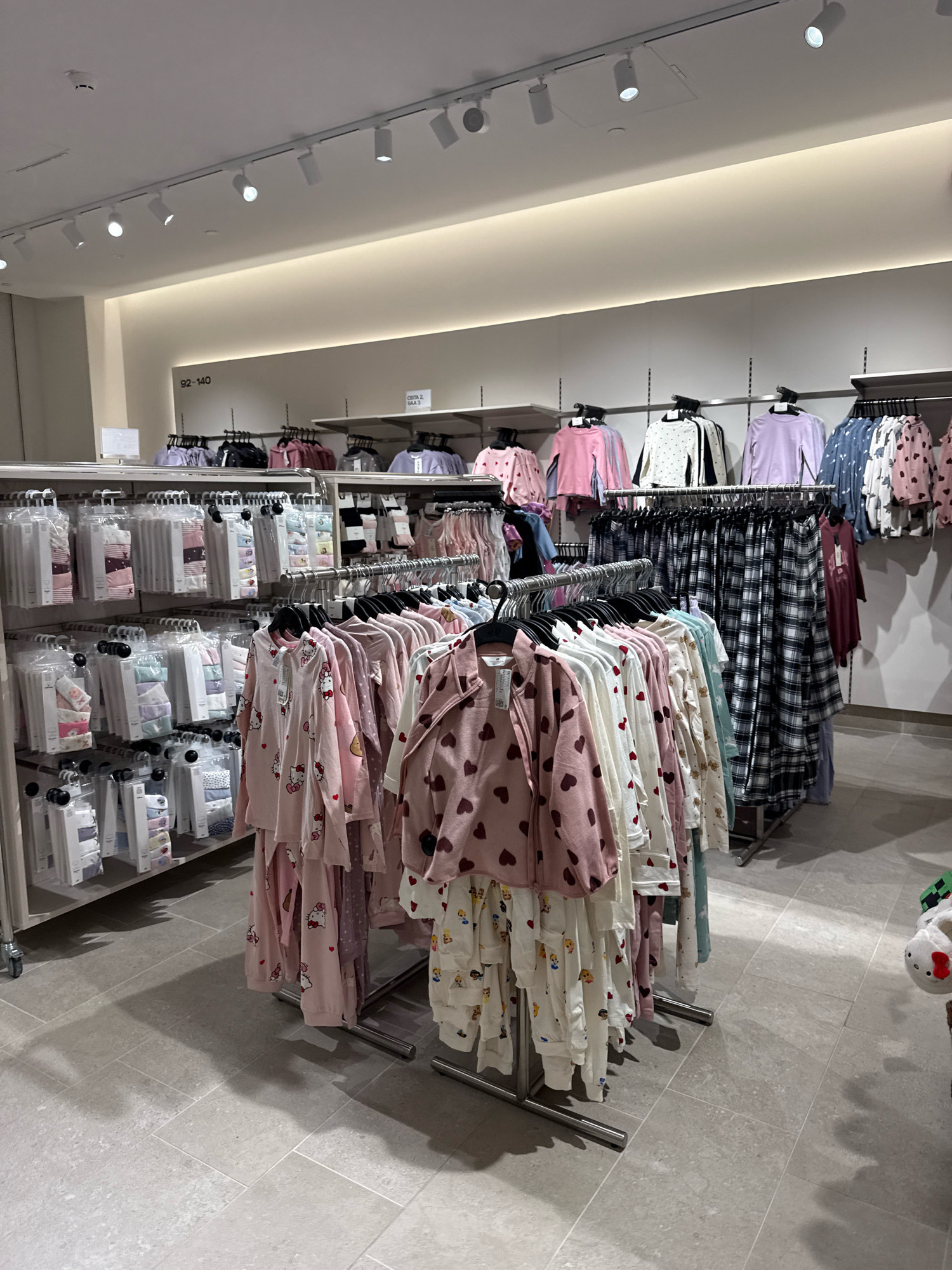 The image shows the interior of a retail store, likely a clothing store, in Tallinn, Estonia. The scene is brightly lit with overhead track lighting. The store is organized with clothing racks displaying various items, primarily children's pajamas and loungewear, some of which are packaged. Several racks are filled with brightly colored sleepwear, some featuring patterns like hearts, polka dots, and cartoon characters such as Hello Kitty. In the background, there are shelves stocked with more packaged clothing. The flooring is tiled with light gray rectangular tiles. The overall impression is one of a well-stocked and organized retail environment. There are no people visible in the image.