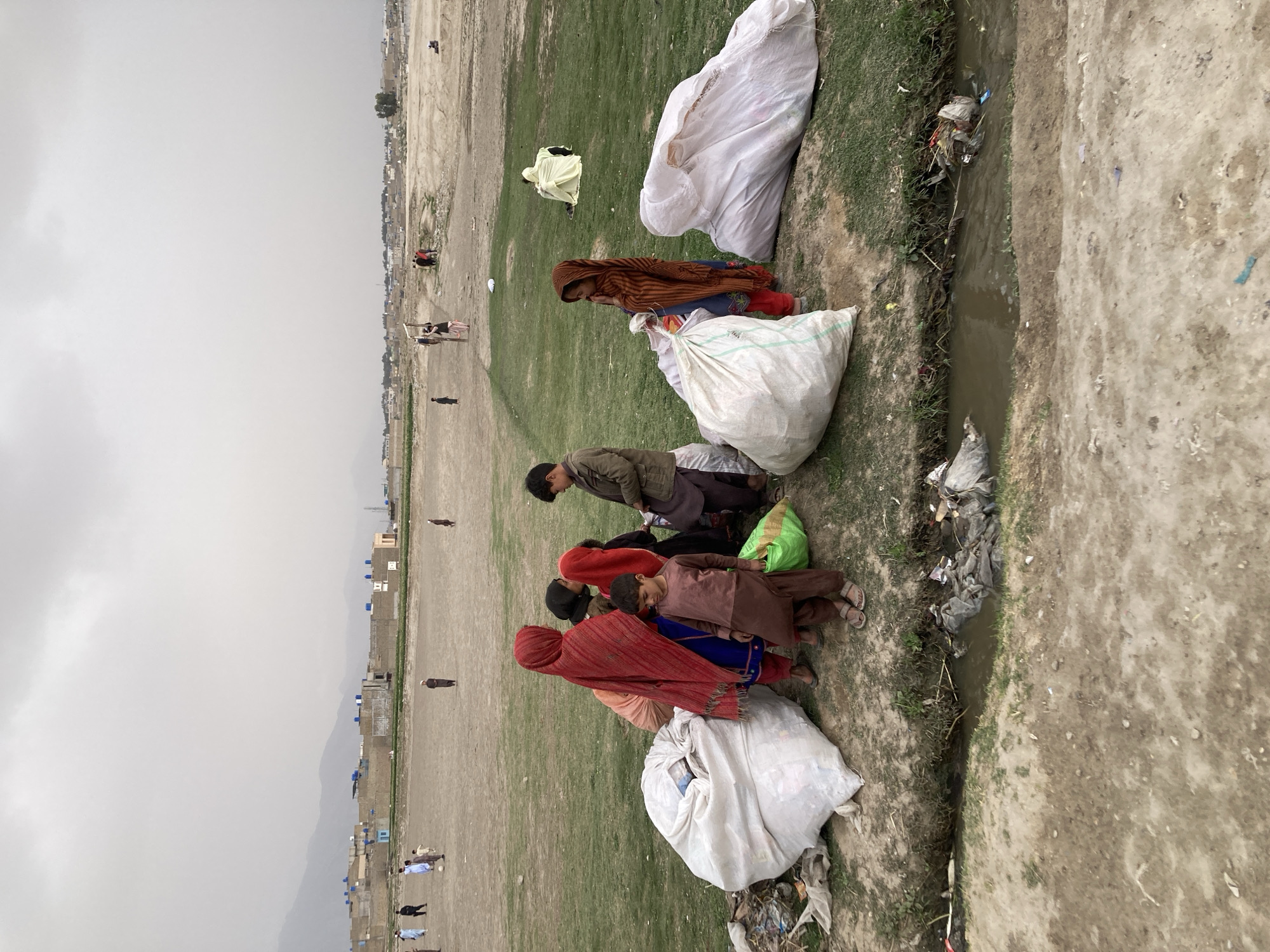 In Quetta City Tehsil, Pakistan, several individuals, including adults and children, are gathered on a grassy verge adjacent to a narrow, trash-filled ditch or stream. Some people are sitting or kneeling near large, white, sack-like bags. The individuals are wearing various clothing, including red headscarves and brown jackets. In the background, a flat, open area stretches toward a distant settlement or city, with buildings visible along the horizon. The sky is overcast.