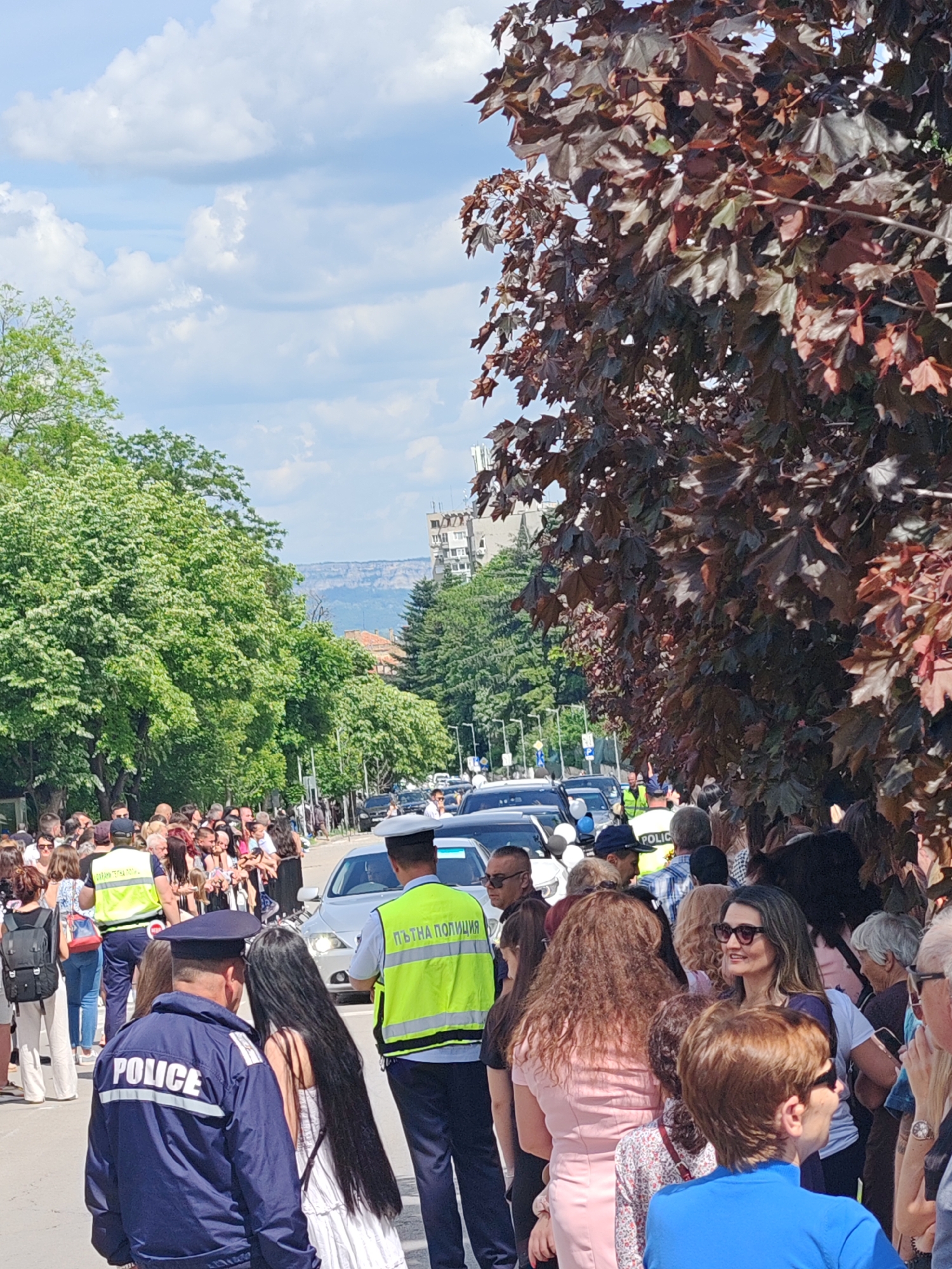 A crowd of people, mostly women, stand on a street in Shumen, Bulgaria. A police officer wearing a navy blue jacket with "POLICE" printed on the back and a peaked cap stands facing away from the viewer, slightly to the left of the frame. To his right is another officer wearing a bright yellow-green vest with "ПЪТНА ПОЛИЦИЯ" printed on the back, also facing away. A line of cars is visible beyond the crowd. Mature green trees are on the left side of the street, and trees with red leaves are on the right. The sky is partially cloudy. A building can be seen further down the street. A mountain range is visible in the distance.