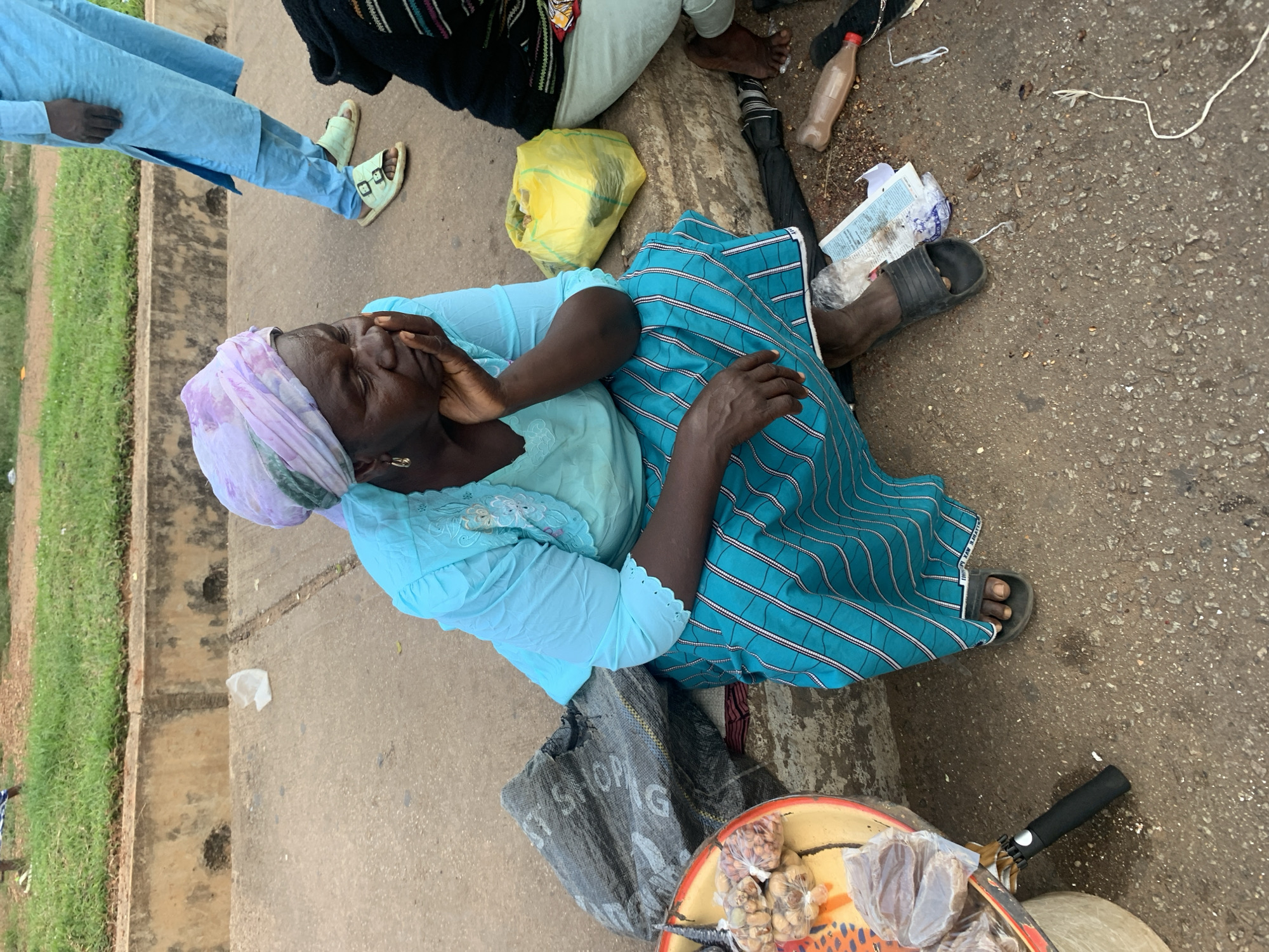 In Barakin Lerawi, Nigeria, an elderly woman sits on a concrete sidewalk, her chin resting on her hand. She wears a light blue blouse with intricate embroidery around the neckline and a blue and white striped skirt. A light purple and white headscarf covers her hair. Beside her, a large black tarp lies on the ground, and to her left, a round tray displays various goods, including small bags of nuts and some wrapped items. A yellow plastic bag is visible behind her. In the background, another person in a light blue outfit stands, and further back, a grassy area is visible beyond a low concrete barrier. The ground is paved with concrete and scattered with debris.