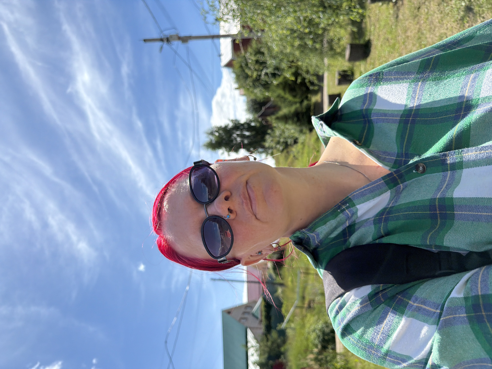 The image shows a person from the shoulders up, taken from a close-up perspective. The person has bright red hair, is wearing dark sunglasses, and has facial piercings, including a nose ring with a turquoise stone and earrings with similar stones. They are also wearing a green and blue plaid shirt. The background consists of a bright blue sky with wispy white clouds, and a blurred natural setting with greenery and what appear to be houses and utility poles. The sunlight suggests it is daytime, likely midday given the brightness. The overall mood seems casual and perhaps relaxed, with the person looking slightly away from the camera. There is no visible text in the image.