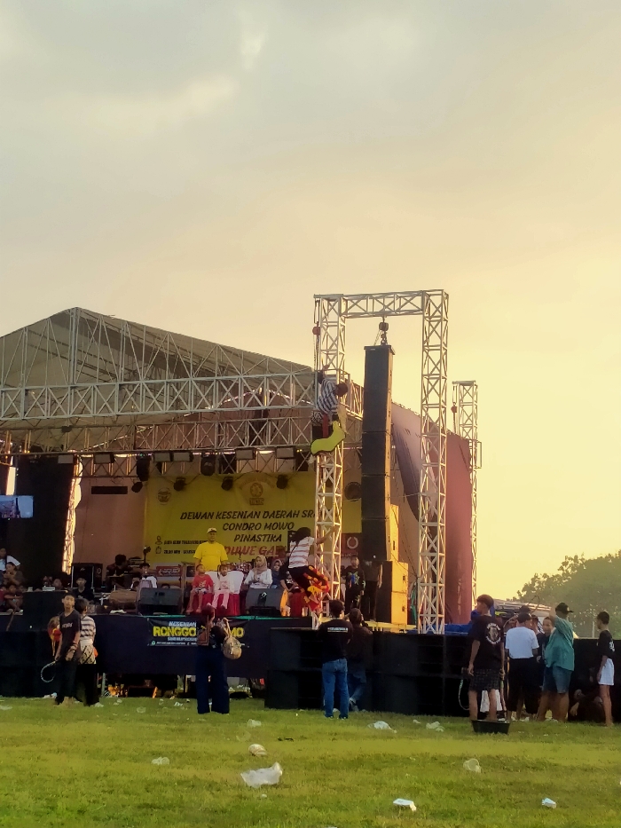The image displays an outdoor public event in Glonggong, Indonesia, observed during a period of warm, golden light suggestive of sunrise or sunset. A large, elevated stage constructed from metal trussing with a roof structure is centrally positioned. A yellow banner across the stage backdrop displays Indonesian text, partially legible as "DEWAN KESENIAN DAERAH SRIRAHAYU CONDRO MOWO PINASTIKA."
On the stage, a man in a prominent yellow shirt is seated. To the right of the stage, a substantial array of professional audio speakers is suspended from a separate metal truss structure. A person in a striped shirt and dark pants is actively suspended from or climbing a yellow, curved structure positioned adjacent to the speaker array. Another individual, also in a striped shirt, is captured in mid-air, performing a jump or dance near the right side of the stage.
The foreground consists of a grassy field, which serves as the audience area. This area is visibly littered with various discarded items, including plastic bags and bottles. Numerous spectators are present, casually dressed in t-shirts, shorts, and jeans, observing the stage. In the immediate foreground, a man in a dark t-shirt and patterned shorts is standing on a dark, bucket-like object.