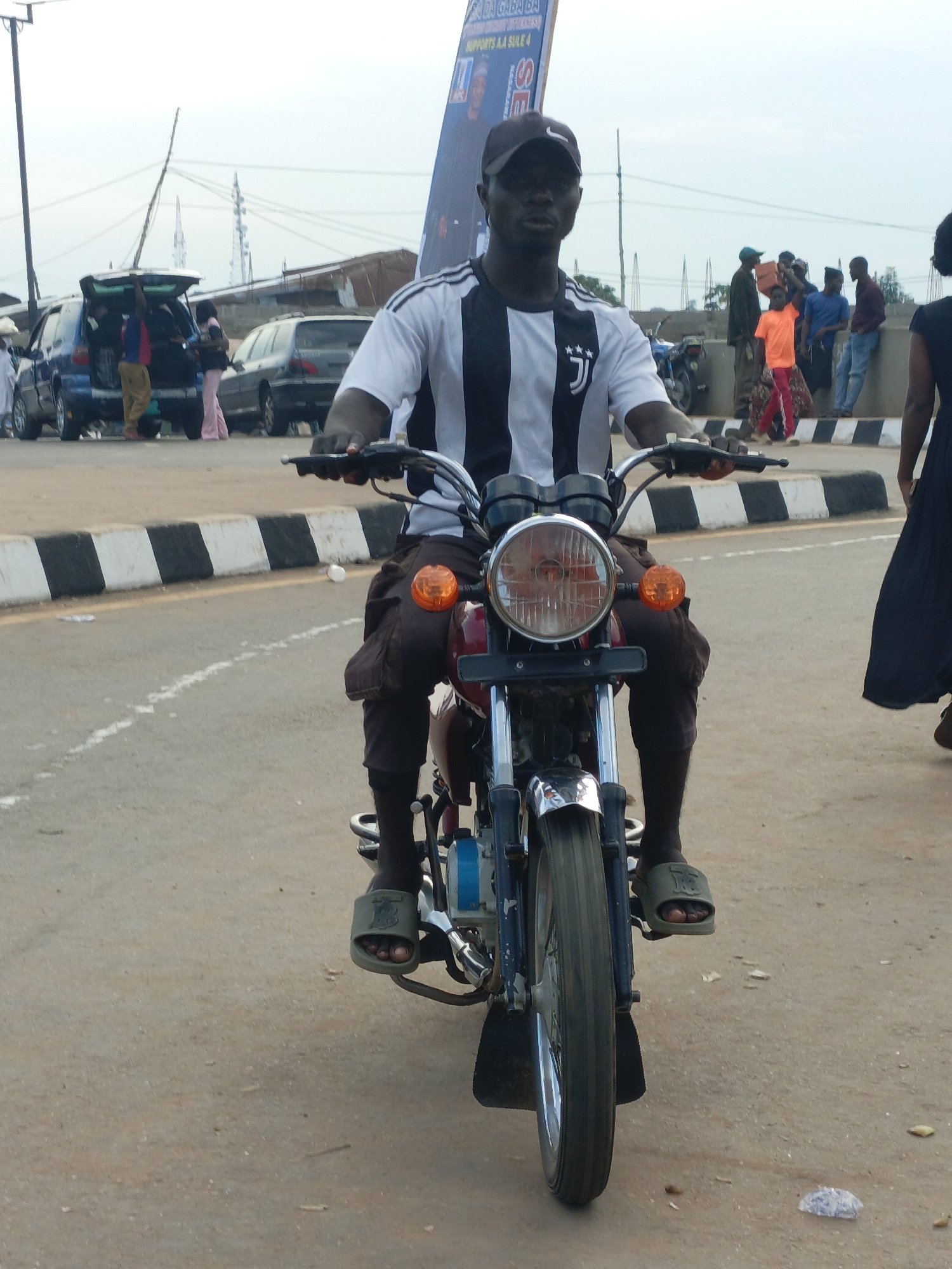 A man is riding a motorcycle in Akwanga, Nigeria. He is wearing a black and white striped shirt and dark pants. His right hand is on the handlebar, and his feet are on the footpegs. The motorcycle is red and blue. In the background, there are several people standing around, some cars, and a large banner. The banner has text on it, but it is not fully legible. The sky is overcast, suggesting it might be late afternoon or early evening. The ground is paved with asphalt and there are some white lines marking the road. The scene appears to be in a public area, possibly a street or a market. The people in the background are dressed in casual clothing. The overall atmosphere seems to be that of a typical day in a bustling Nigerian town.