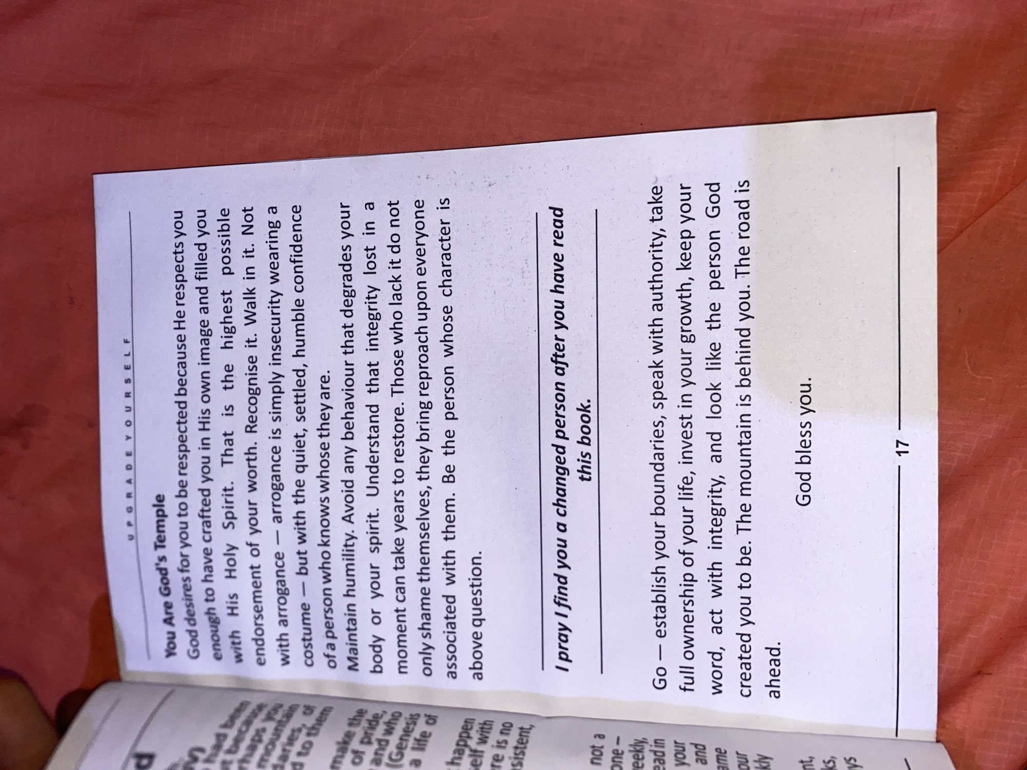 The image shows an open book with printed text. The page on the right is mostly visible and includes text under the heading "You Are God's Temple" and subheadings such as "I pray I find you a changed person after you have read this book". The page number "17" is visible at the bottom right corner. The book is resting on a surface with a reddish hue. The book appears to be lying horizontally with a slight tilt. The location is not identifiable in the image.