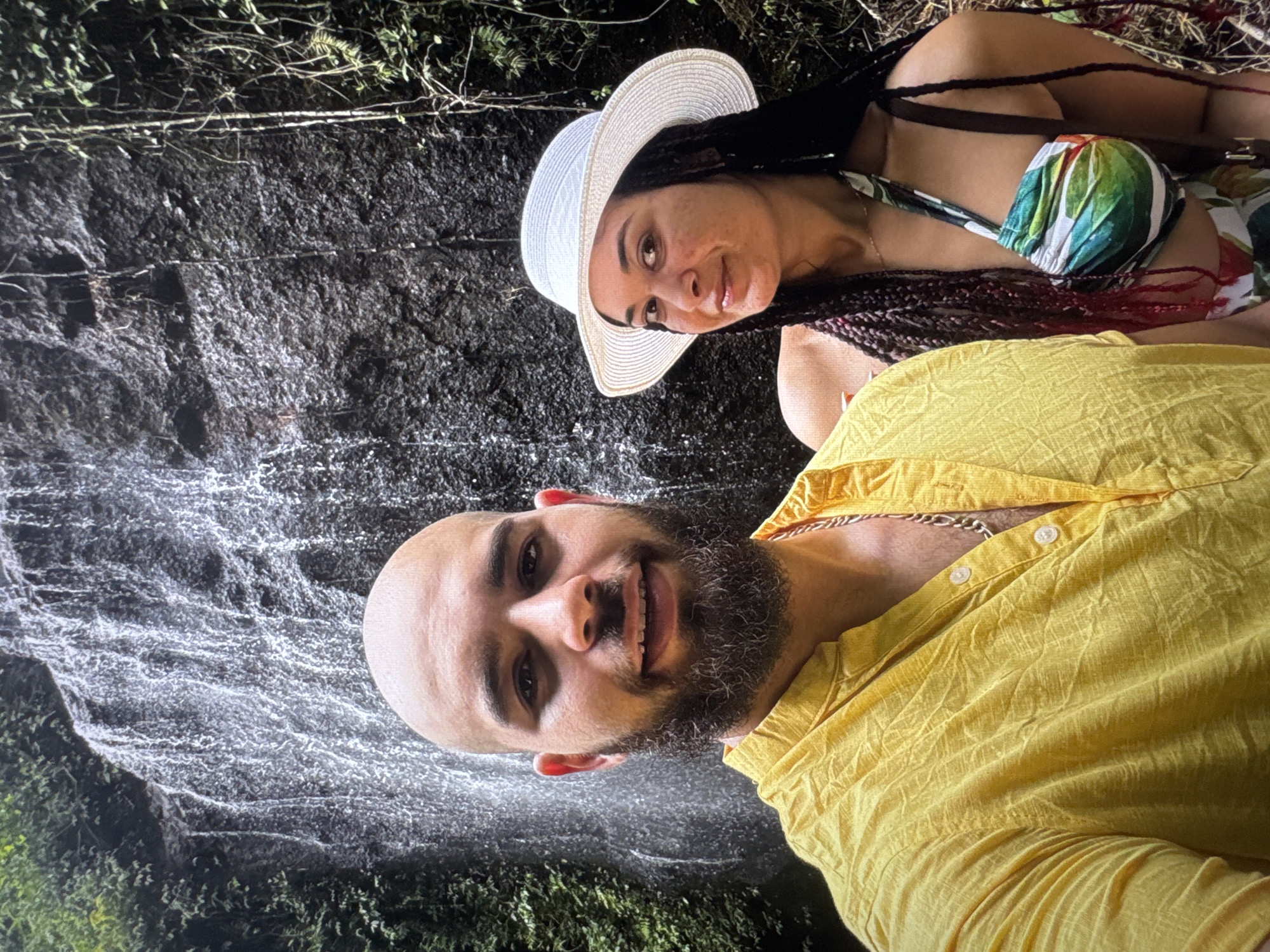 The image shows a man and a woman posing for a photo in front of a waterfall in an unknown location. The man is bald with a beard, wearing a yellow shirt and a silver necklace. The woman is wearing a white sun hat and a floral bikini top. The waterfall is in the background with surrounding green vegetation.