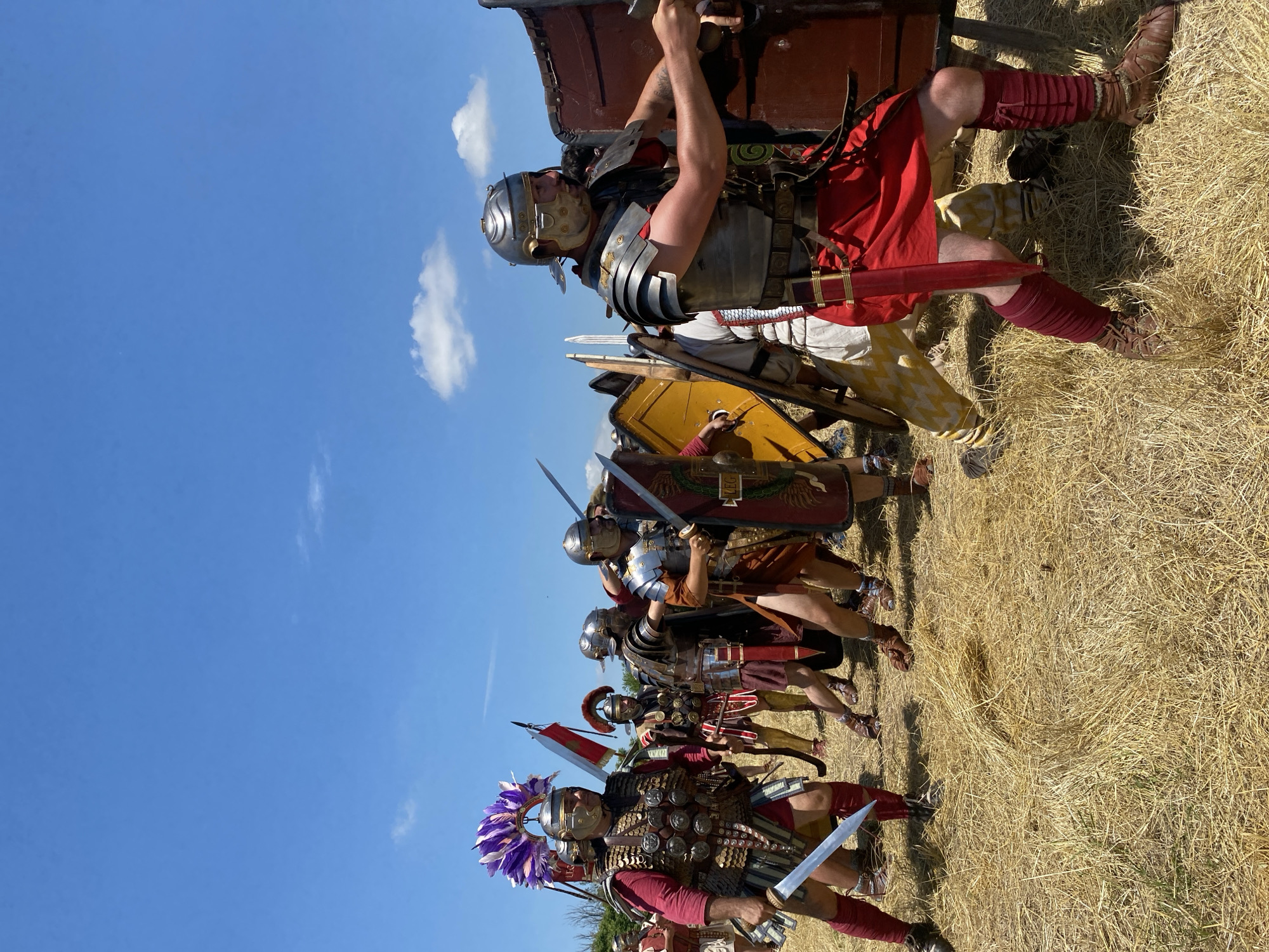 The image captures a historical reenactment of Roman soldiers engaged in a formation outdoors under a clear blue sky with scattered white clouds. The scene is set in a dry, golden-brown field of straw or stubble, located in Gigen, Bulgaria.
Approximately eight individuals, costumed as Roman legionaries, are visible in the foreground and mid-ground. They are equipped with replica Roman military gear, including segmented plate armor (lorica segmentata), ornate helmets with cheek guards, red tunics, and red or brown leg wraps with period-appropriate footwear. Each soldier carries a gladius (short sword) and a scutum (rectangular shield).
The prominent figure on the far left, likely representing a centurion, wears a distinctive breastplate adorned with medallions and a tall purple and white plumed crest on his helmet. He holds his gladius in his right hand, poised. Behind him, a red and white vexillum (military standard) is partially visible.
The soldiers are positioned in a close formation, with several holding their shields forward in a defensive stance. The soldier on the far right actively braces a large, reddish-brown rectangular shield with both hands, displaying the letters "LEG" and an ornate wreath design. Another soldier to the left has a yellow shield with similar markings. Their postures suggest movement or readiness for combat, with swords drawn or held for immediate use. The overall impression is a staged military maneuver or display.