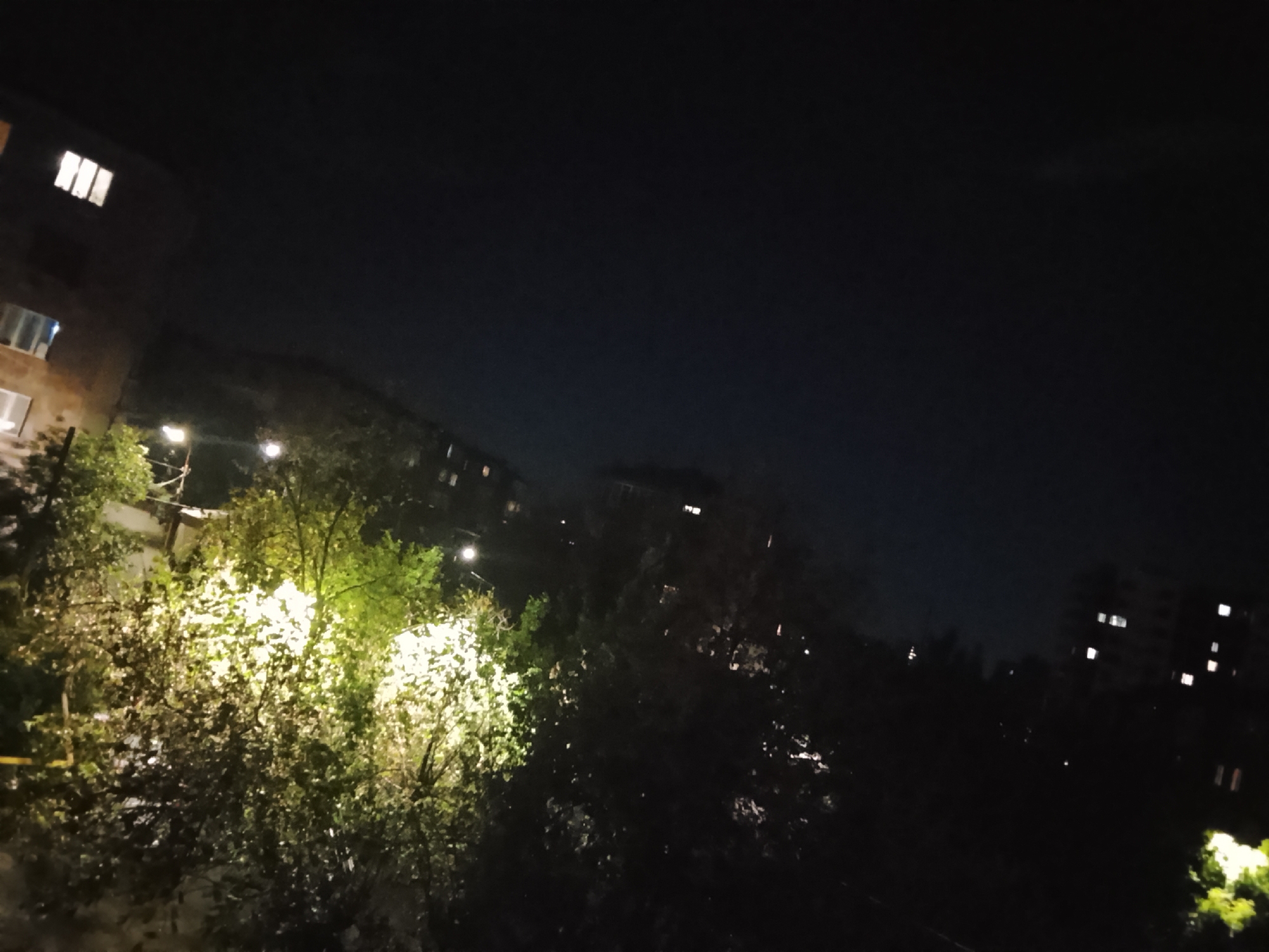 This media file captures a nighttime view of an urban or suburban landscape in Yerevan, Armenia, likely from an elevated perspective such as a window or balcony. The upper portion of the image is dominated by a deep, dark night sky, indicating clear weather.
The scene below the horizon is characterized by a mix of buildings and lush foliage, brightly illuminated by artificial light sources. On the left, a multi-story building with a reddish-brown facade displays several brightly lit windows. Throughout the middle ground, dense trees and bushes are vividly illuminated by streetlights and other unseen light sources, creating bright patches of green and yellow foliage that stand out against the darkness. Several streetlights are visible, notably one on the left and another further right, casting their glow onto the surrounding greenery.
In the distance, the outlines of more buildings can be discerned, marked by scattered, glowing windows. The overall impression is one of a quiet residential area after dark, with artificial lighting creating strong contrasts and highlighting specific elements within the nocturnal setting. No people or specific activities are visible, and there is no discernible text in the image.